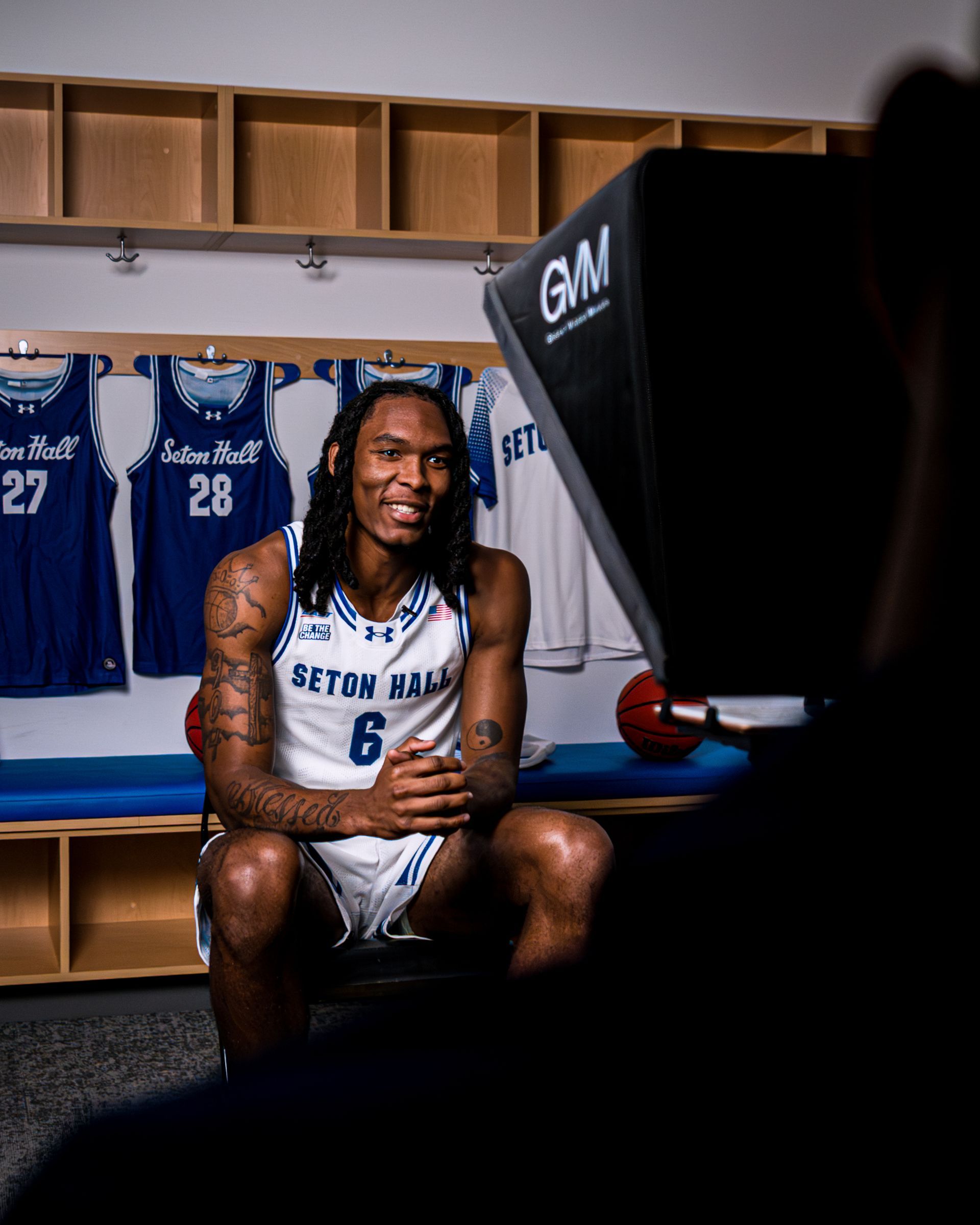 Athlete in white jersey sitting in a locker room, smiling, with blue team uniforms hanging behind him.