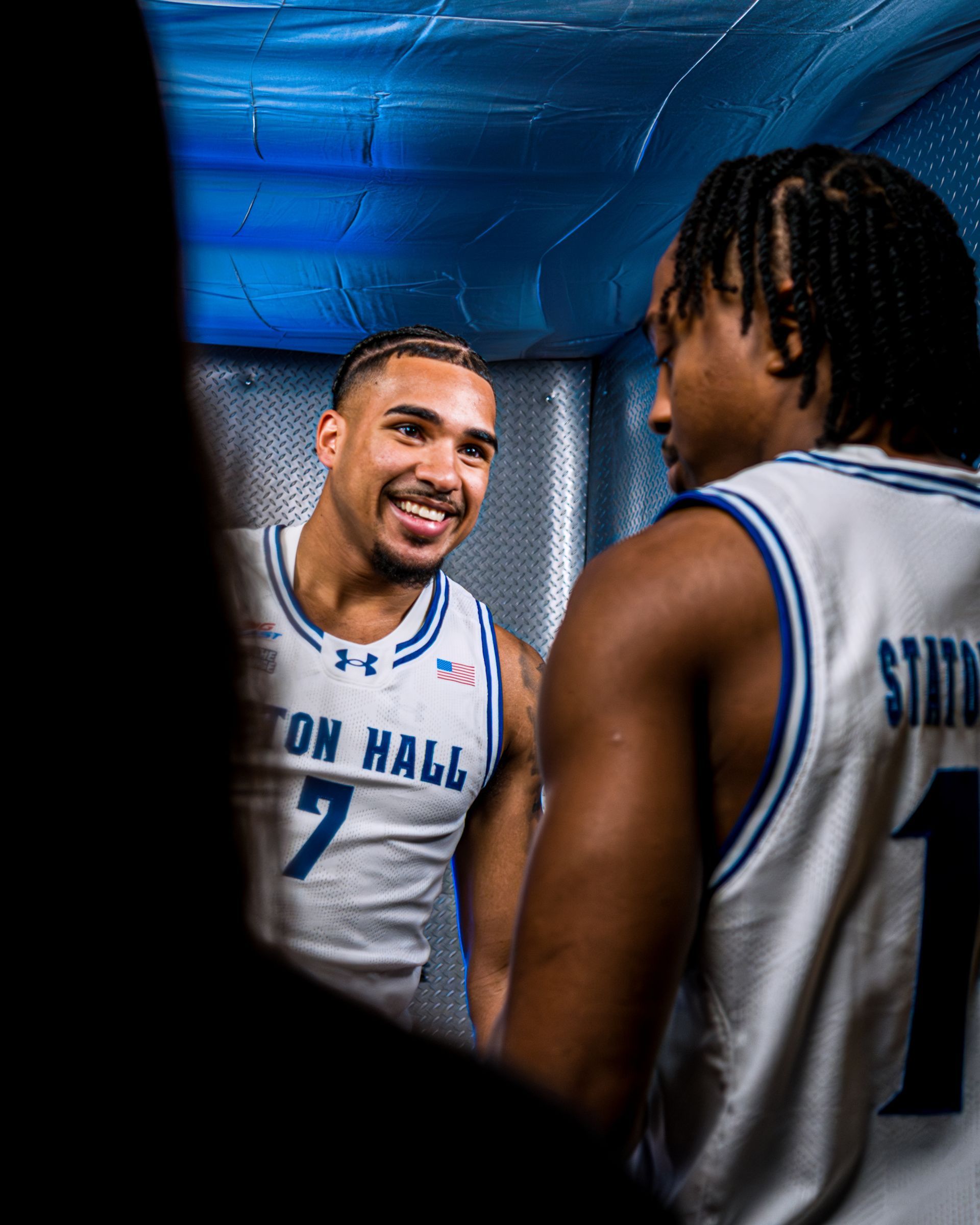 Seton Hall basketball players talking in the tunnel, one smiling in a white jersey with number 7