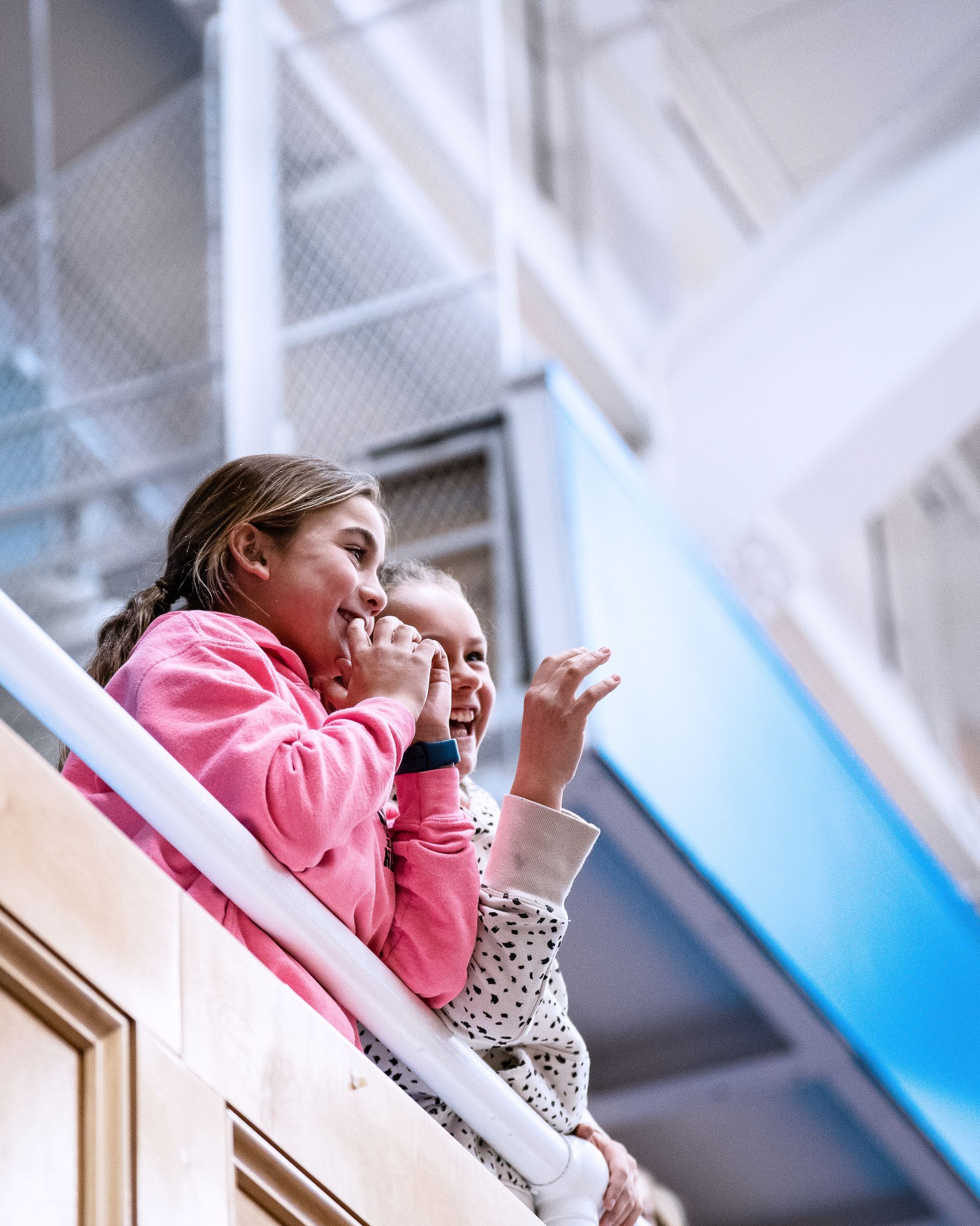 Two children in pink jackets looking down from a balcony indoors