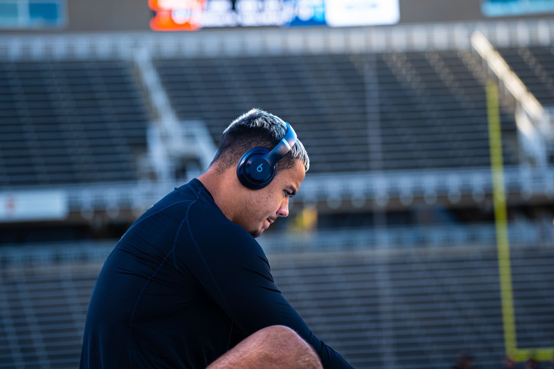 Man in navy shirt wearing blue headphones sits on a stadium bench, looking down.