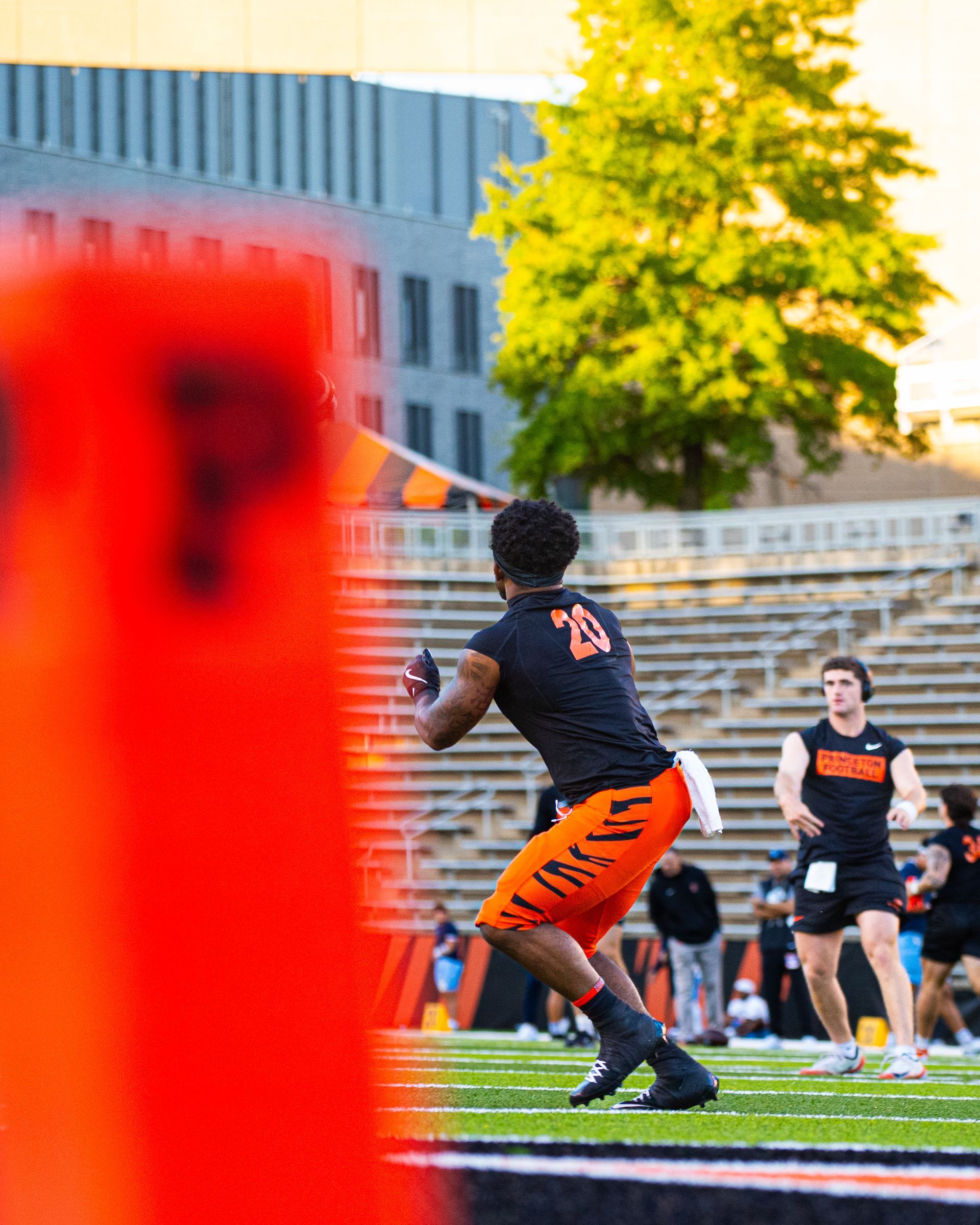 Football player in black and orange runs on field, with blurred orange foreground and stadium steps behind.