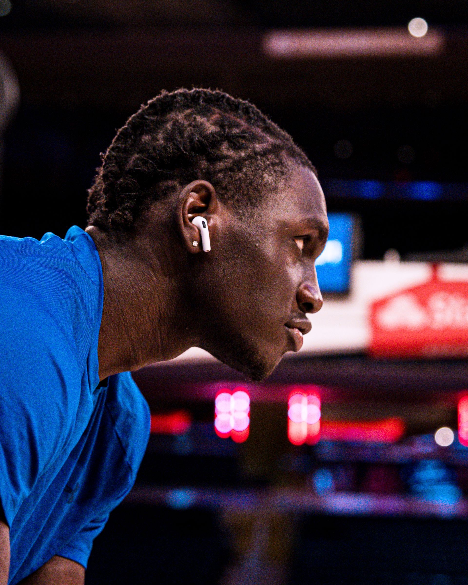 Athlete in blue shirt leaning forward in profile on a sports court, with blurred scoreboard lights behind.