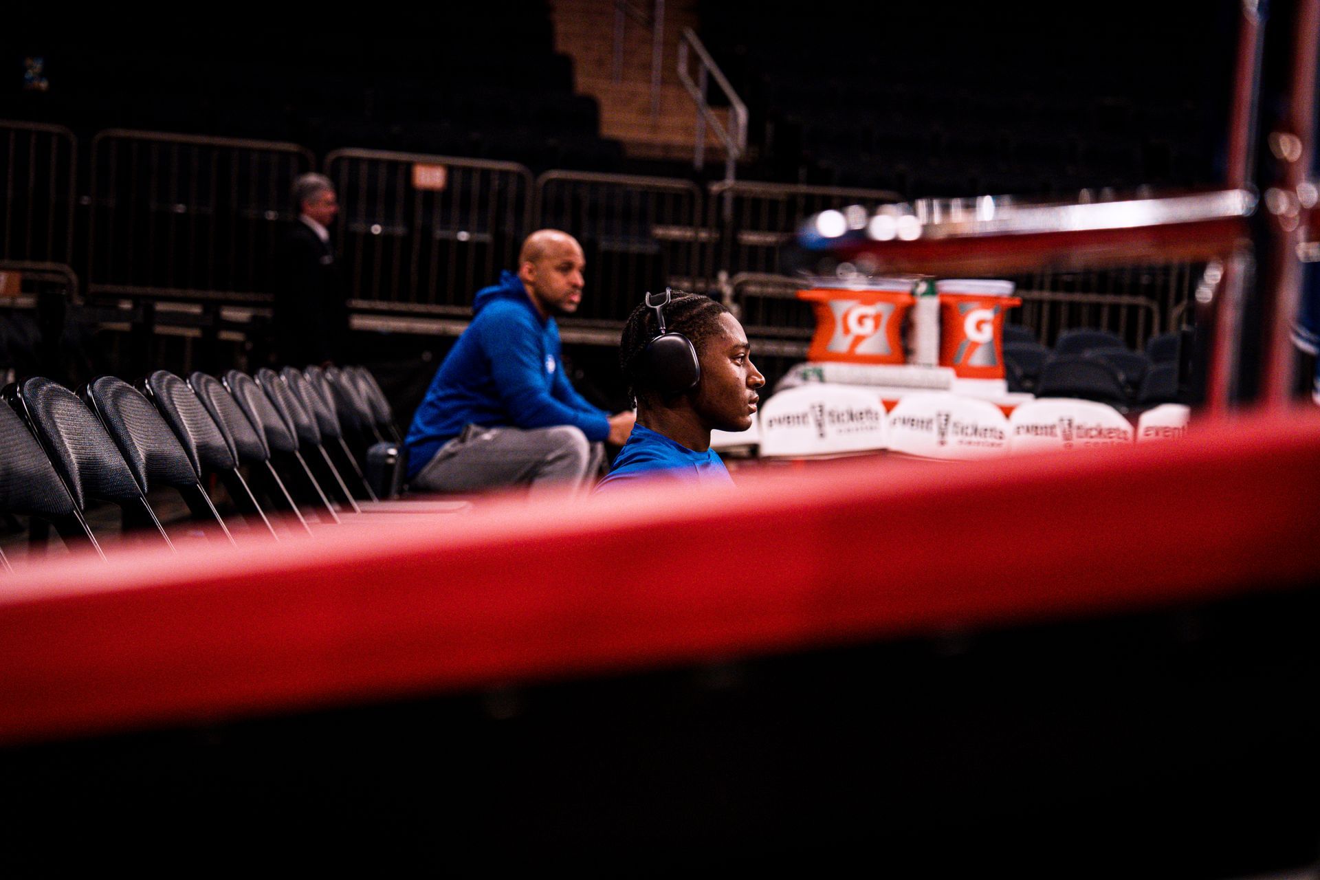 Two people in blue sit ringside in a dim arena, with red ropes and white coolers in the foreground
