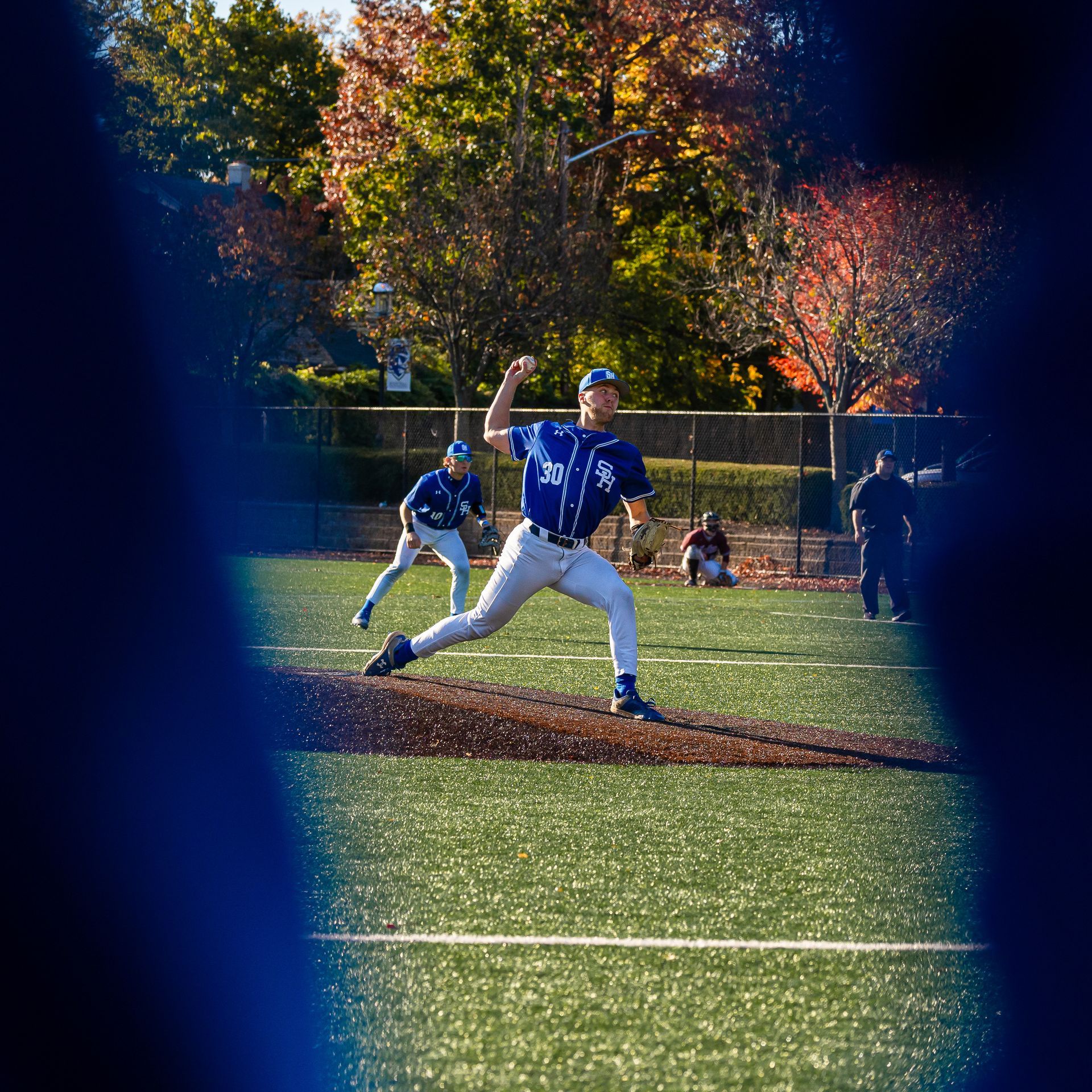 Baseball pitcher in blue winds up on a sunlit field with a runner on base and autumn trees behind him
