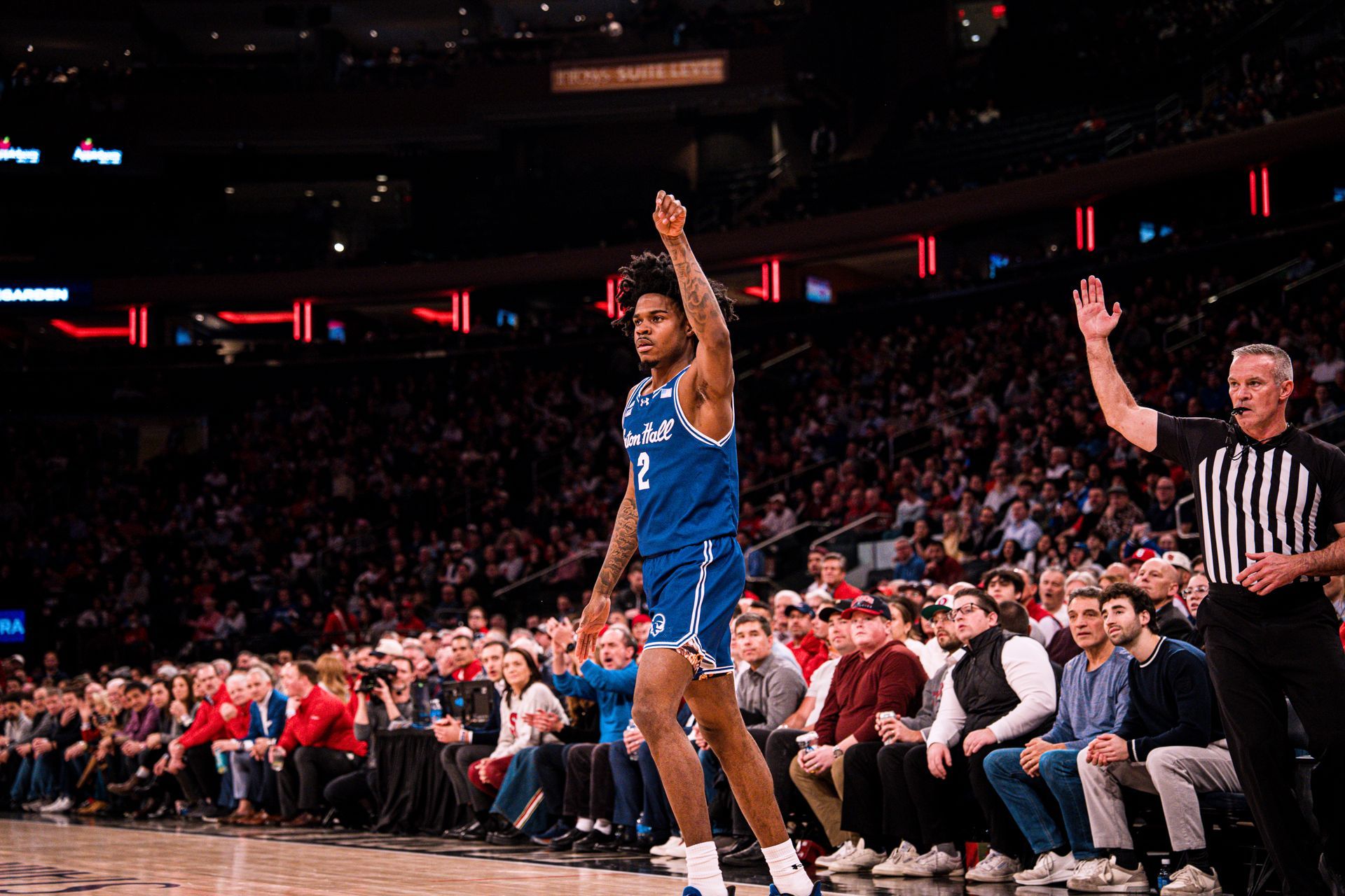 Basketball player in blue jersey raises one finger on court as referee stands nearby and crowd watches