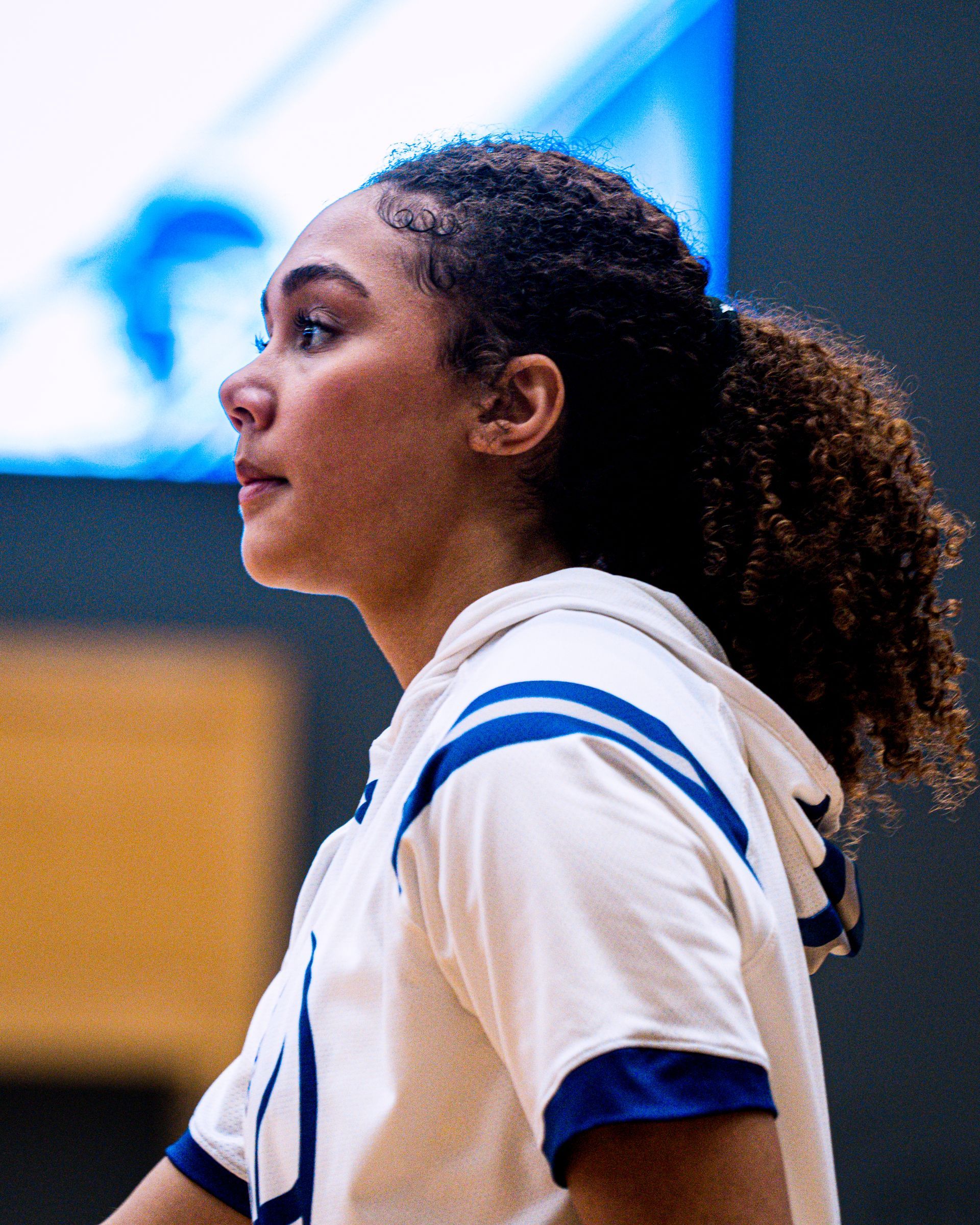 Side view of a person in a white-and-blue jersey standing indoors on a gym court, looking left.