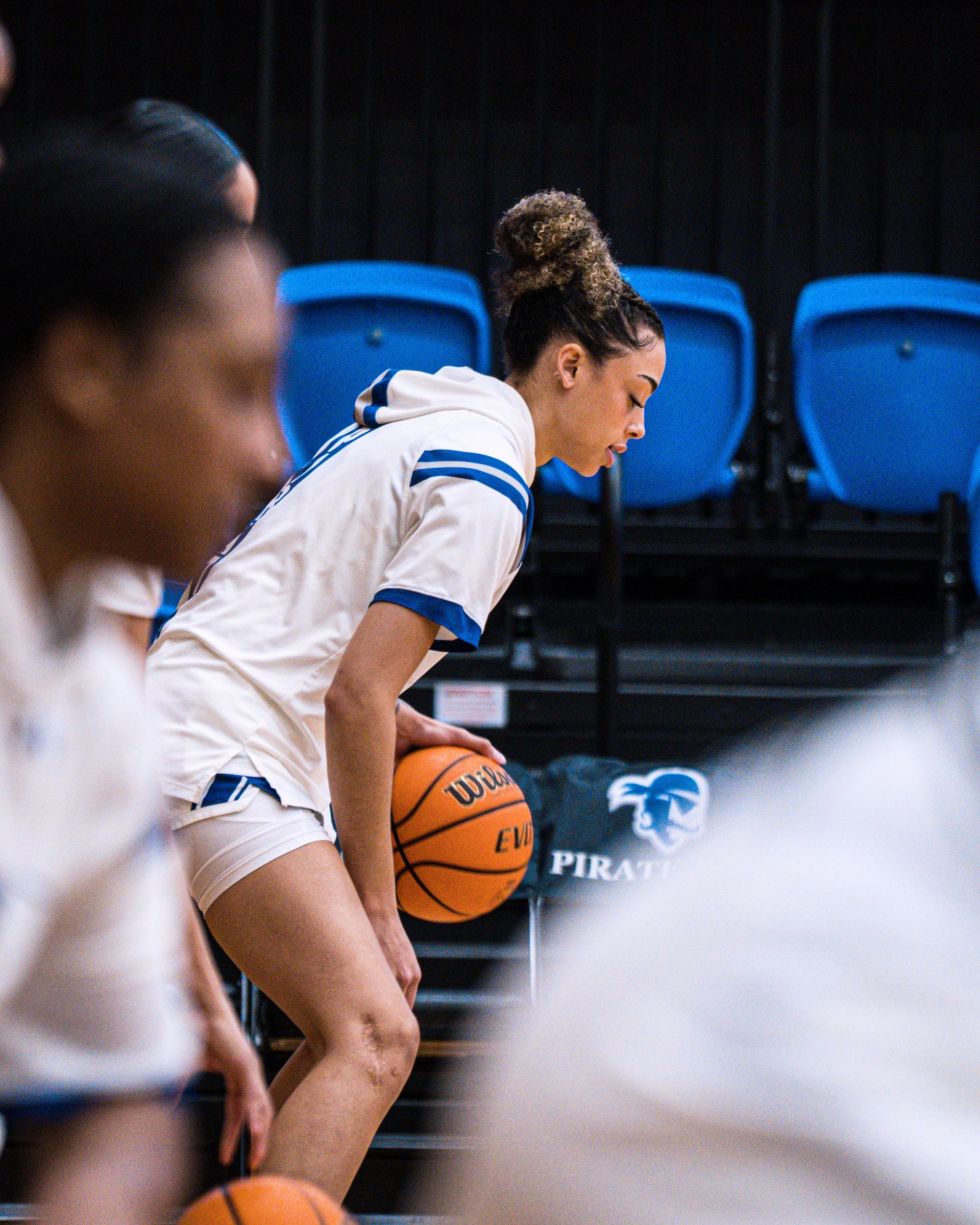 Basketball player in white jersey dribbling on court with blue seats in the background