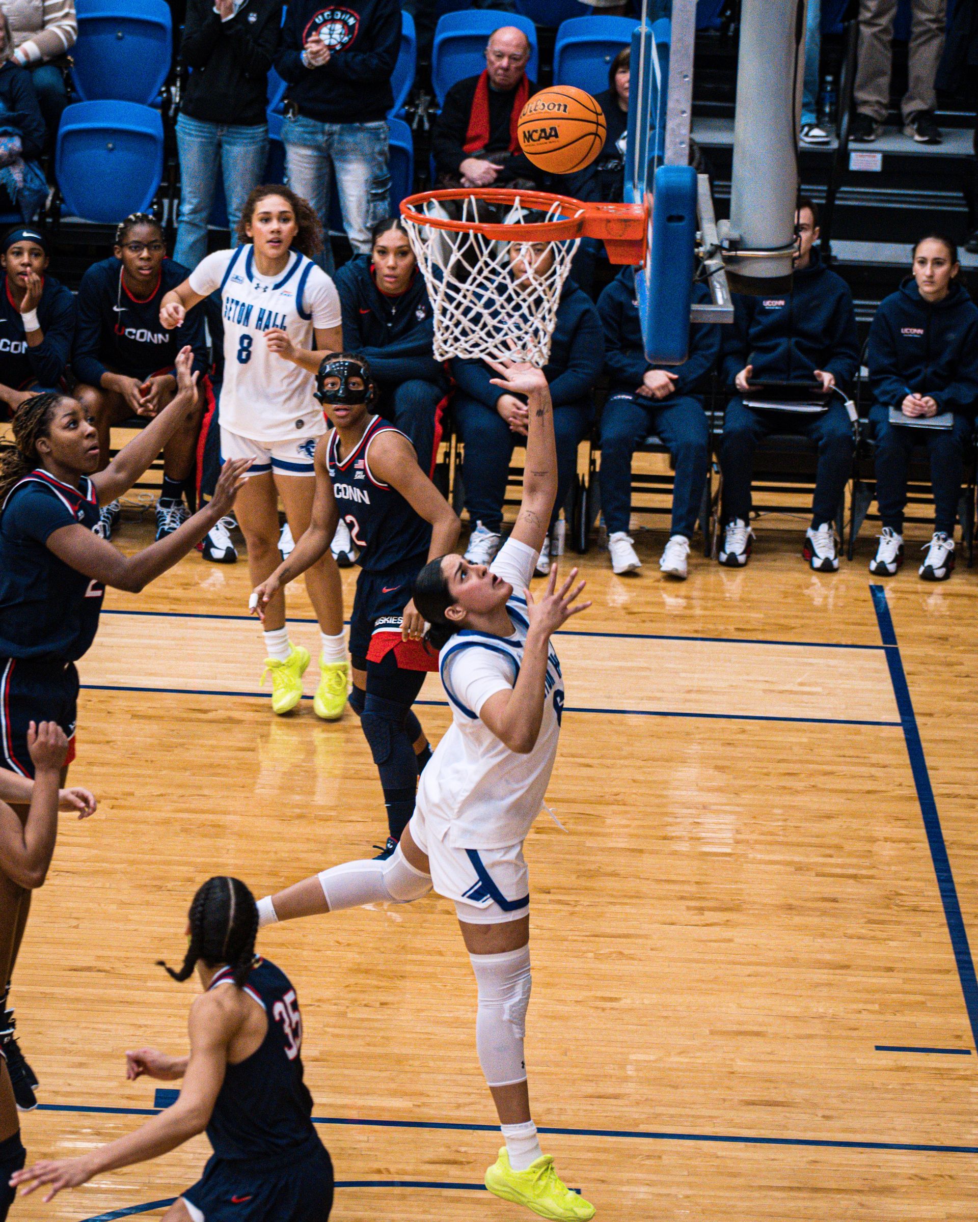 Basketball player in white attempts a layup as defenders and spectators watch indoors.