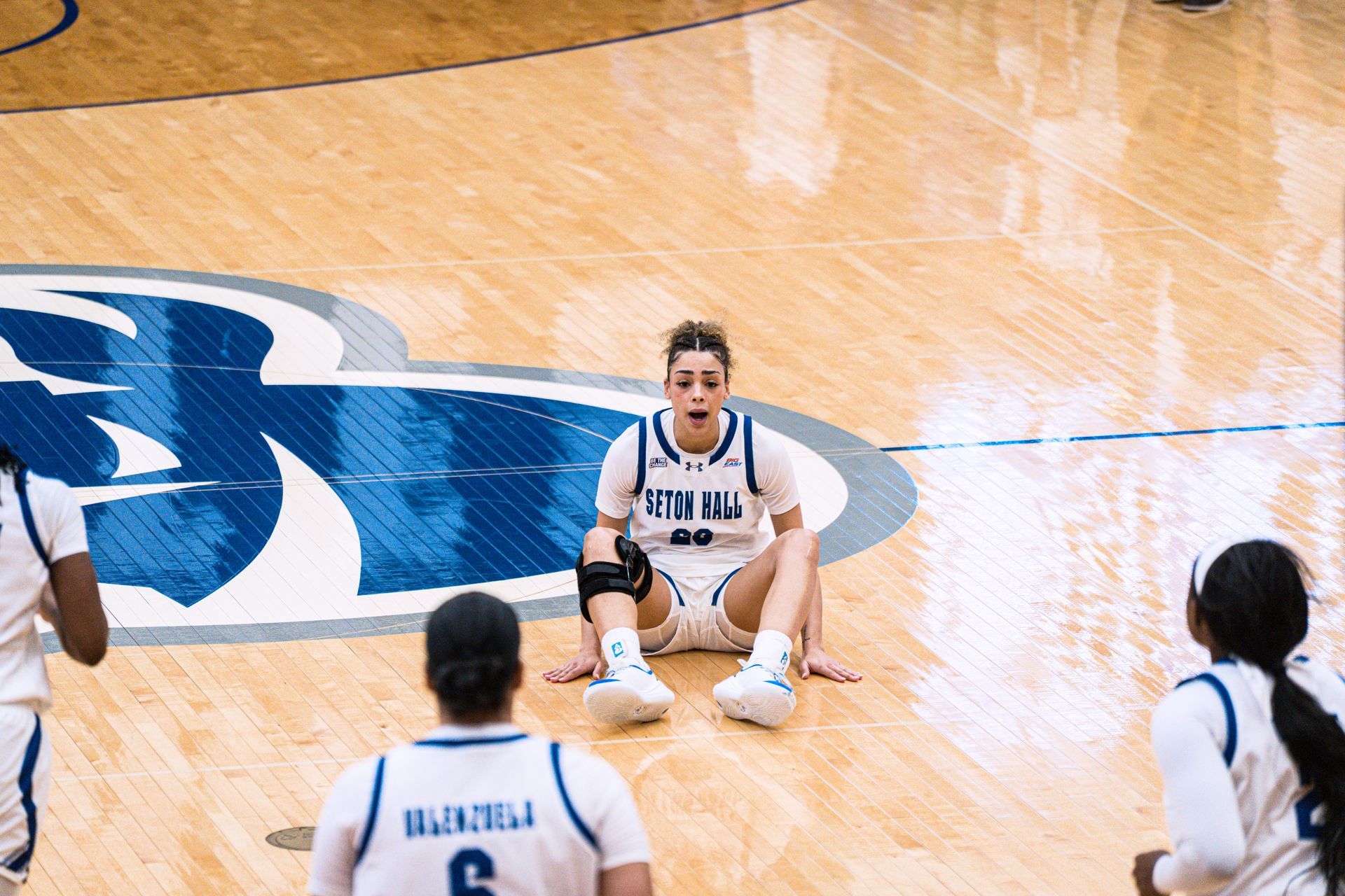 Basketball player sitting on the court near the Duke logo, surrounded by teammates during a game