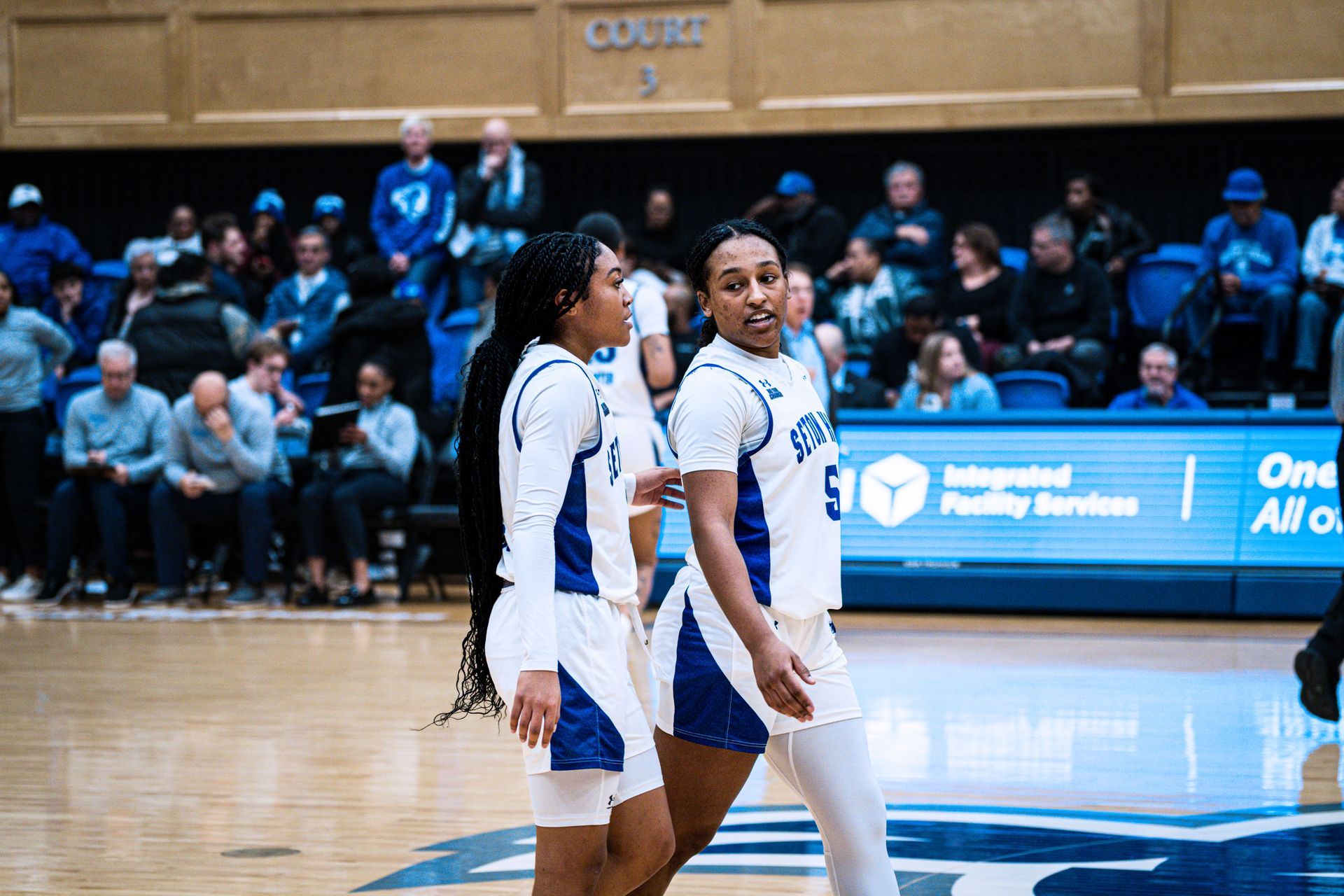 Two basketball players in white and blue uniforms on an indoor court with spectators in the background