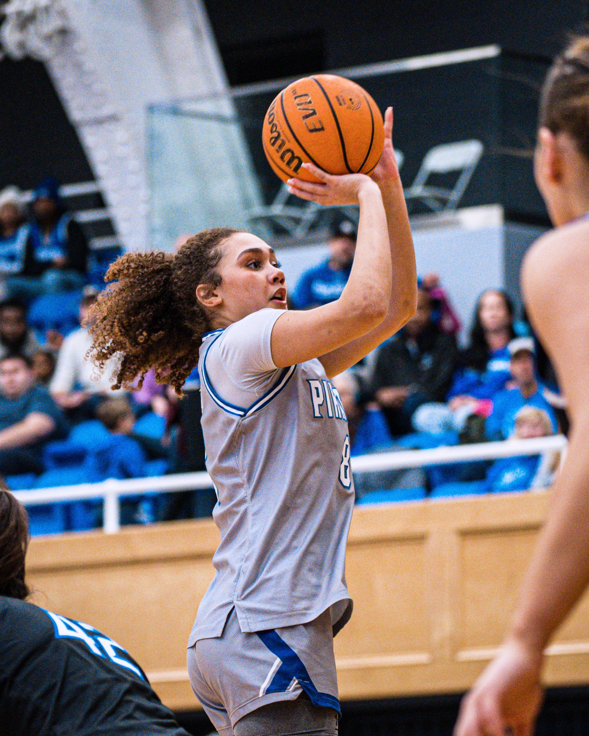 Basketball player in gray jersey shooting the ball in an indoor gym, crowd watching behind her