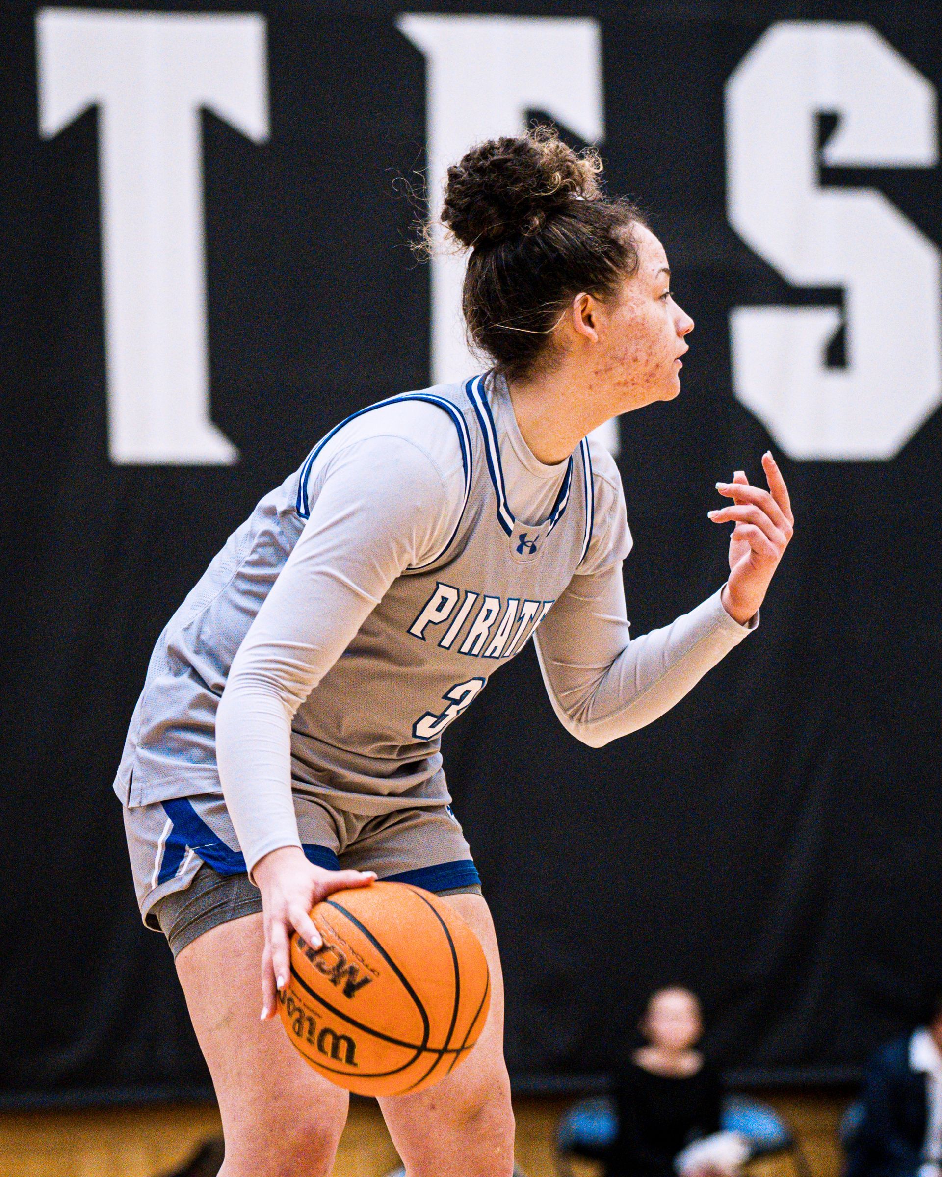 Basketball player in gray and blue dribbles on court, with black wall and large white letters behind her