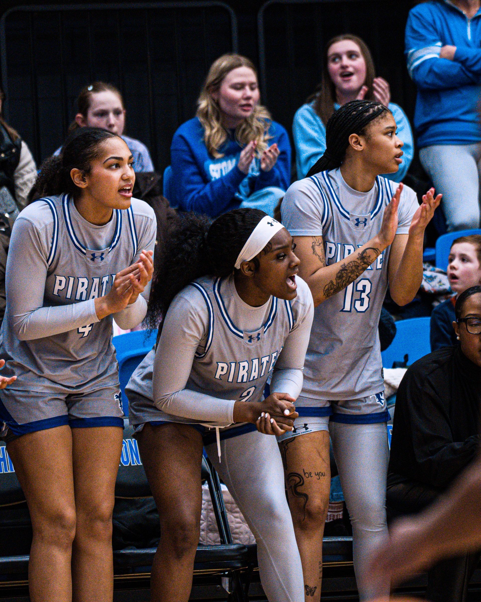 Three women in gray basketball uniforms crouch on the bench, clapping and cheering at a gym game.
