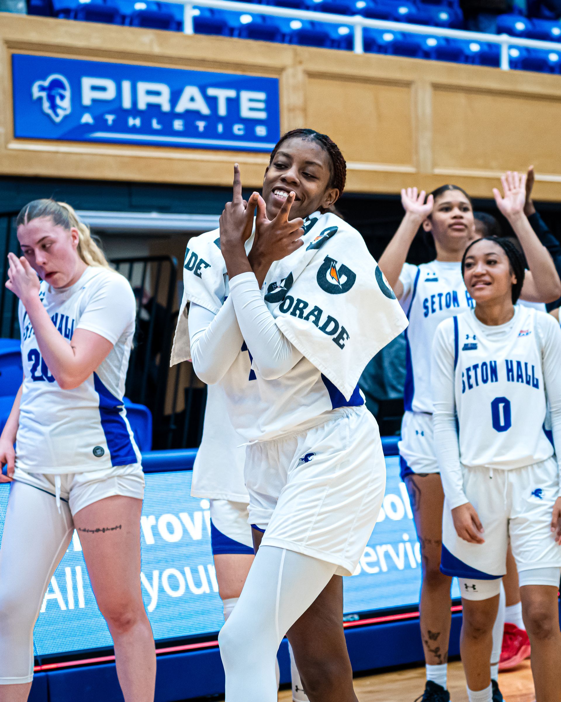 Cheering volleyball players in white uniforms on the court with a PIRATE ATHLETICS sign behind them