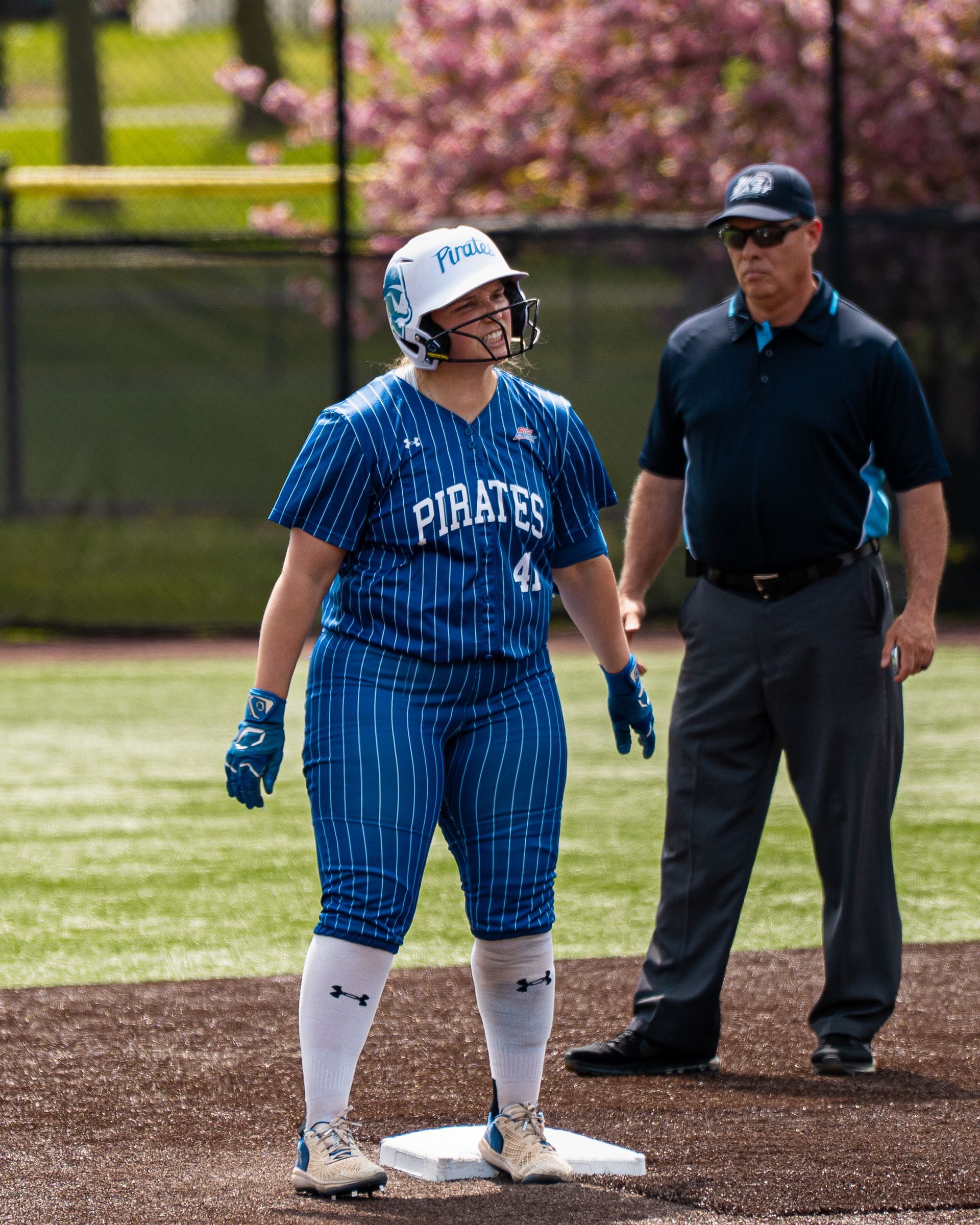 Softball player in blue uniform stands on base while an umpire watches on the field.