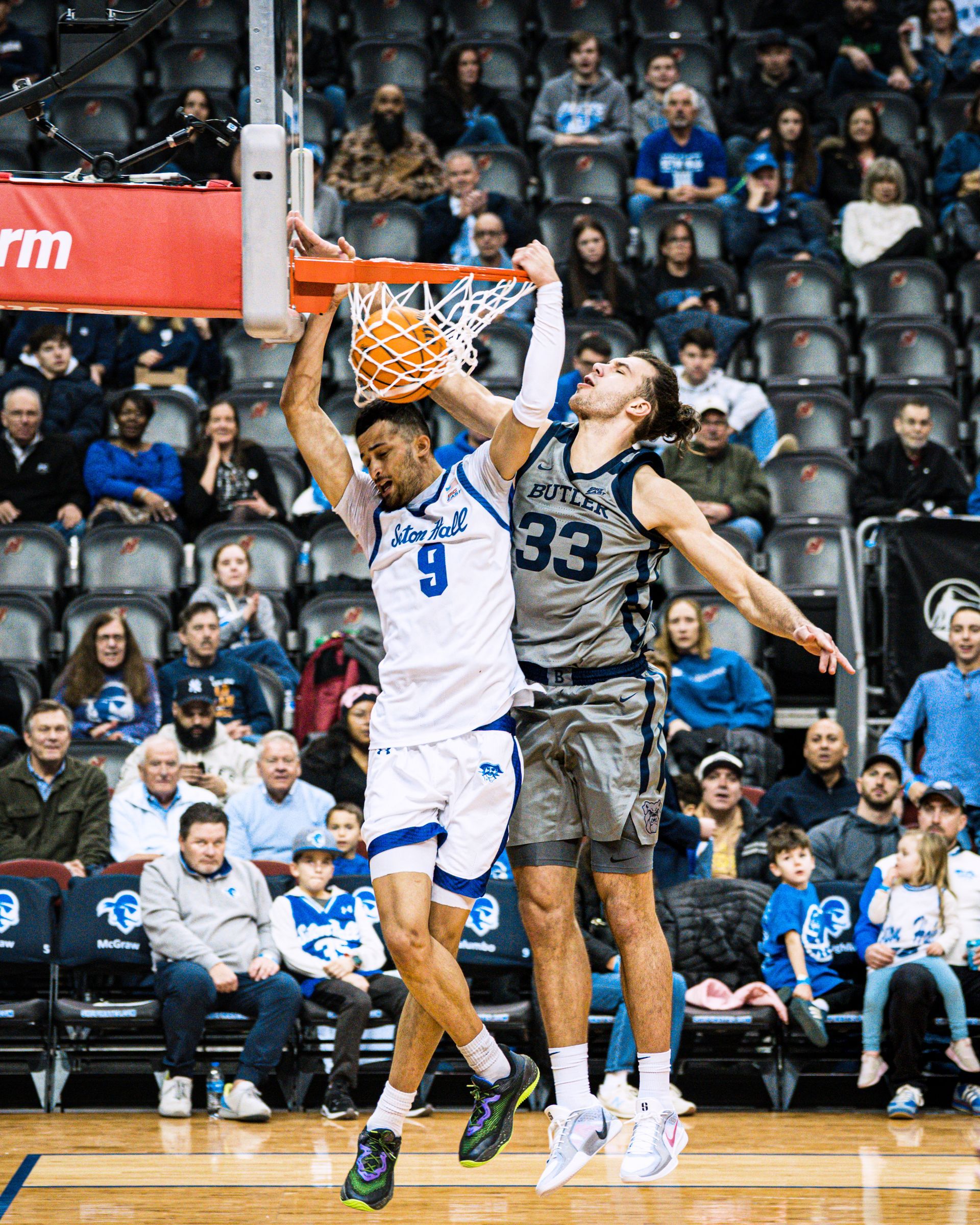 Basketball player in white jersey dunks over defender under the hoop in a crowded arena