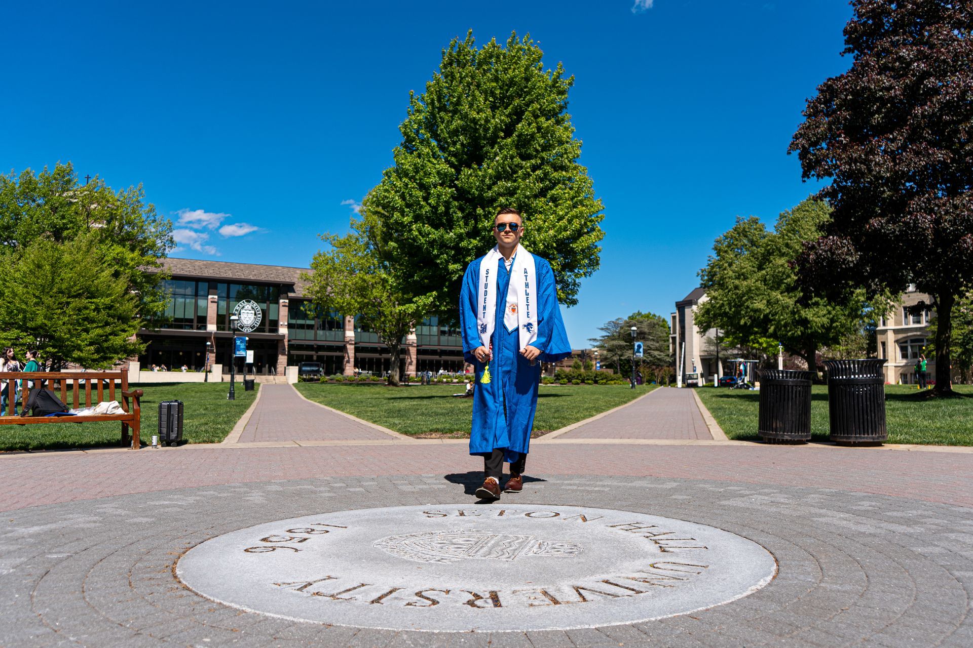 Graduate in blue cap and gown standing on a campus walkway under a bright blue sky