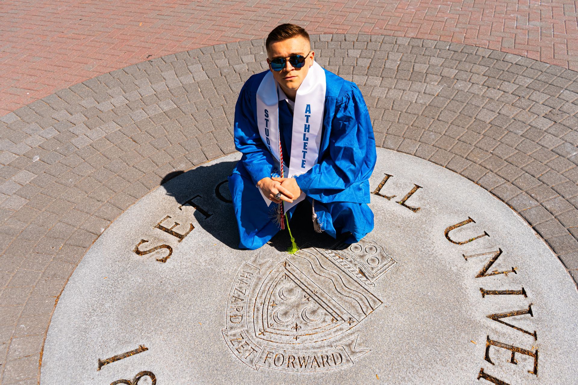 Person in blue graduation gown kneeling on a university seal in bright sunlight, wearing sunglasses.