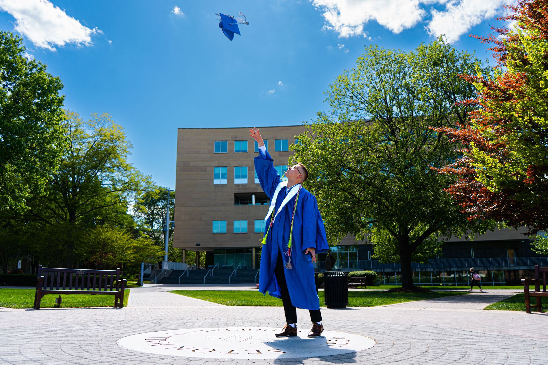 Graduate in blue gown and cap stands in a sunny campus courtyard, raising a diploma against a clear sky.