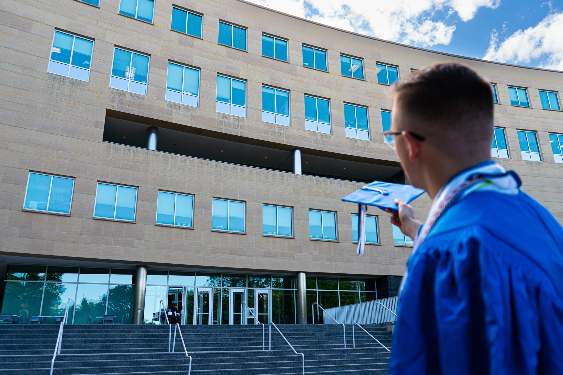 Graduate in blue gown pointing at a modern building with steps and glass entrance