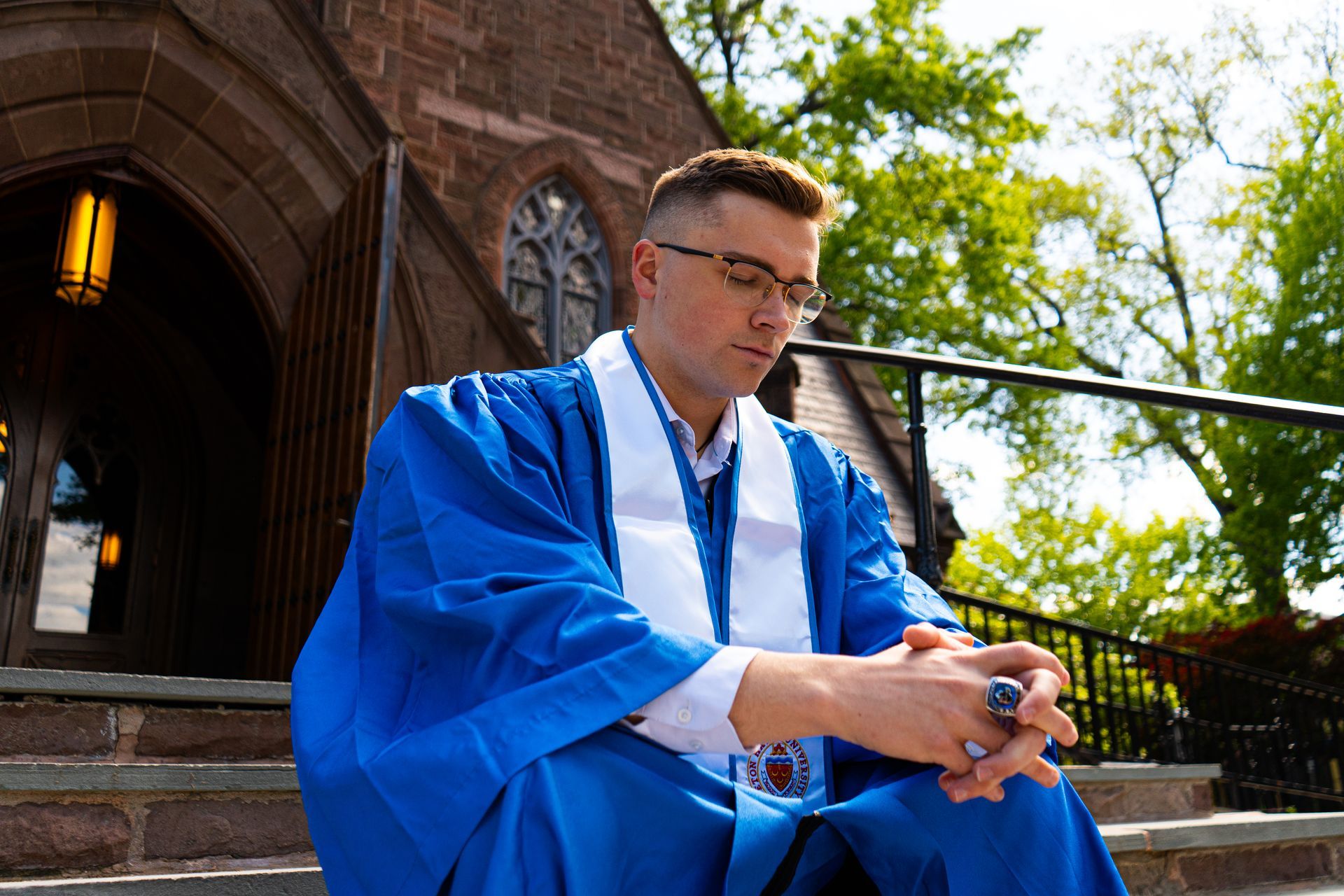 Person in a blue robe sitting on steps outside a brick building, looking down and holding a rosary.