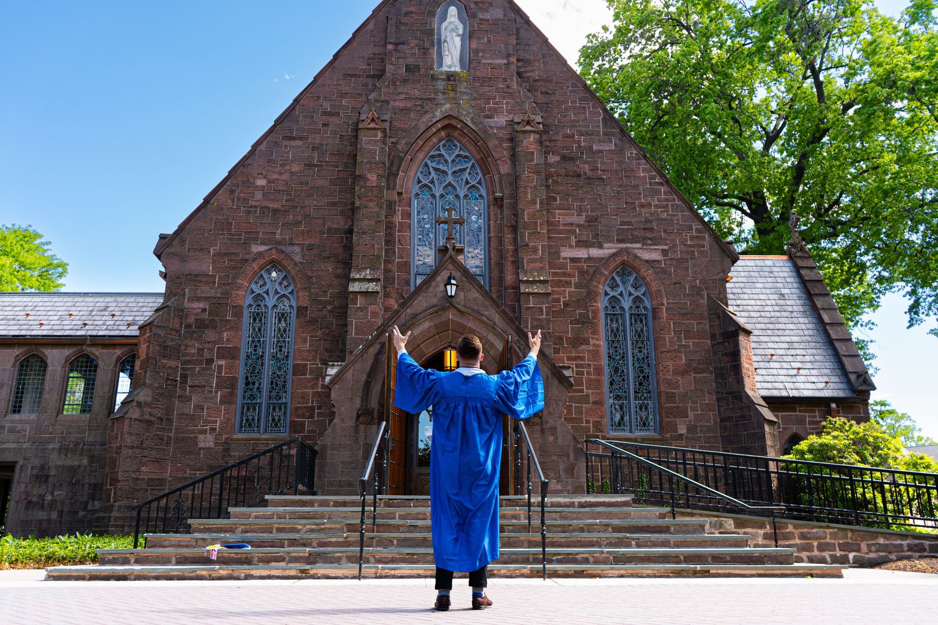 Person in blue gown stands with arms raised before a stone church entrance
