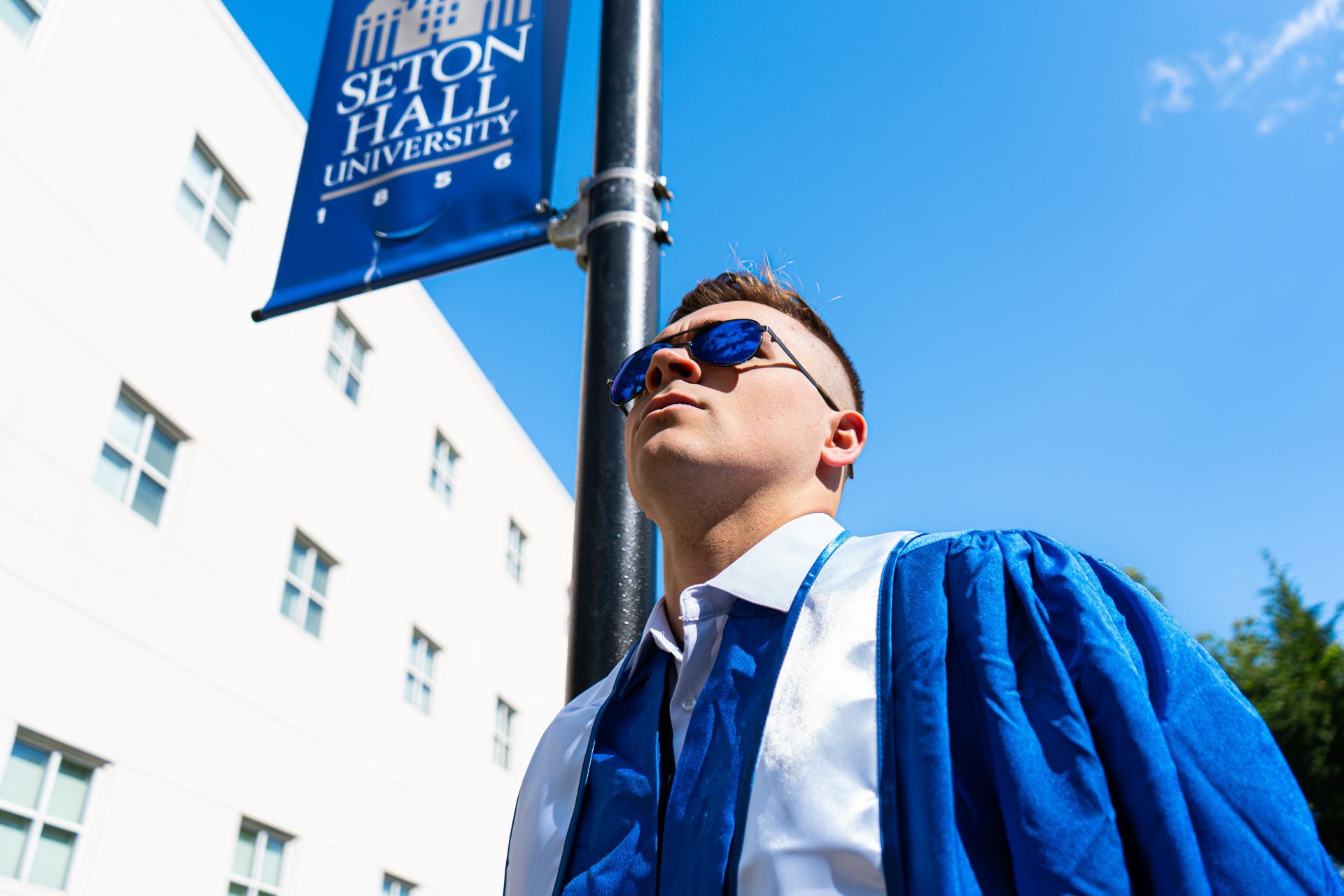 Graduate in blue gown standing by a “Station” banner, looking upward in bright sunlight