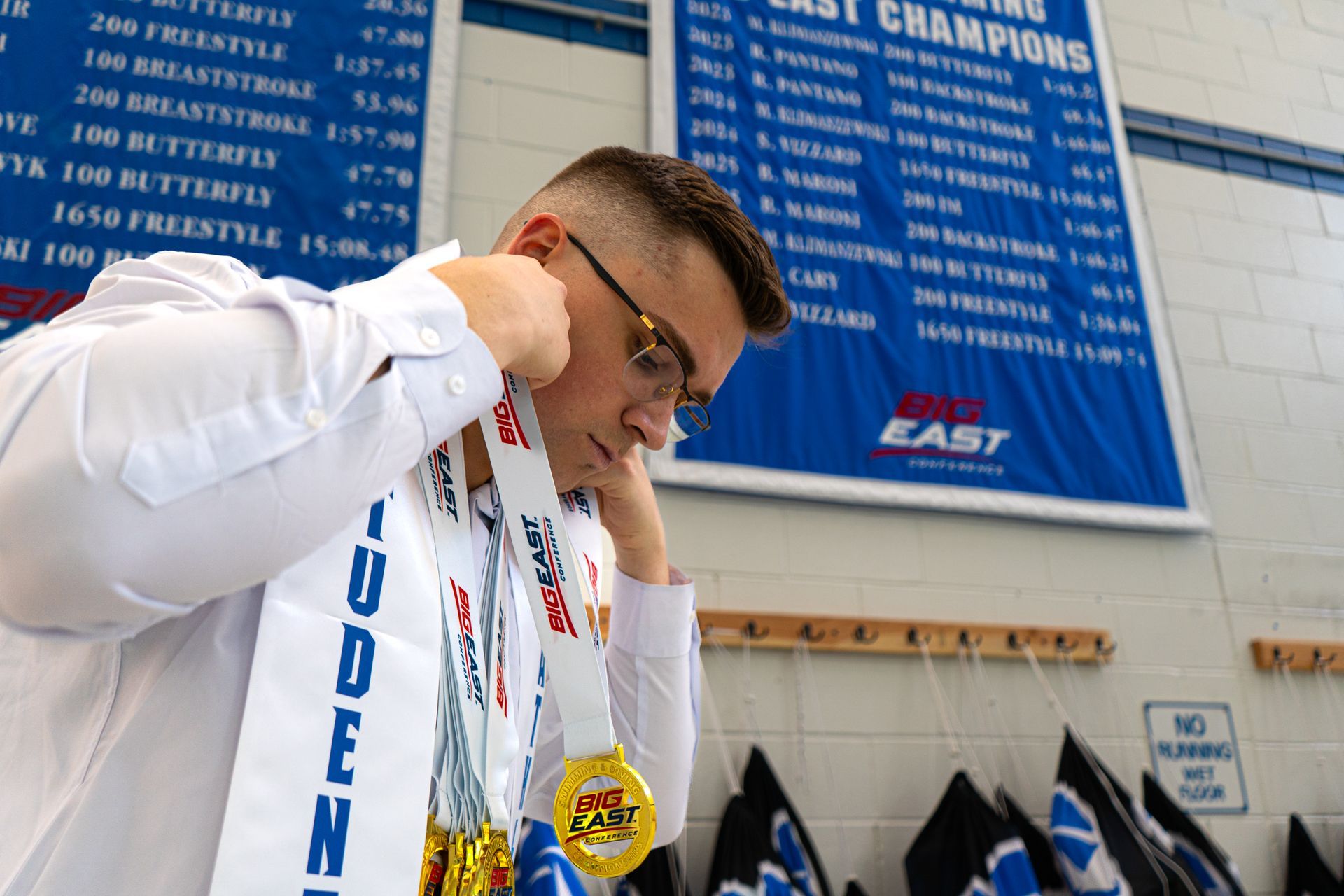 Man wearing medals in a gym, looking down while holding them up beside a championship board
