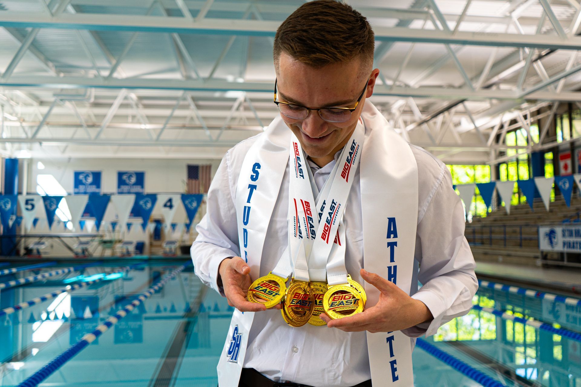 Swimmer holding two gold medals at an indoor pool, looking down and smiling