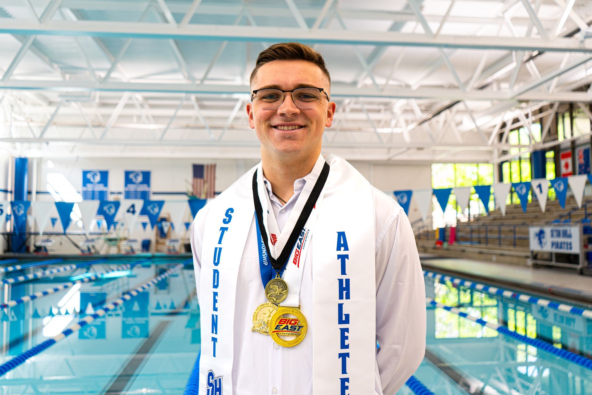 Man smiling in a swim arena, wearing a medal and white towel, with lanes and blue banners behind him