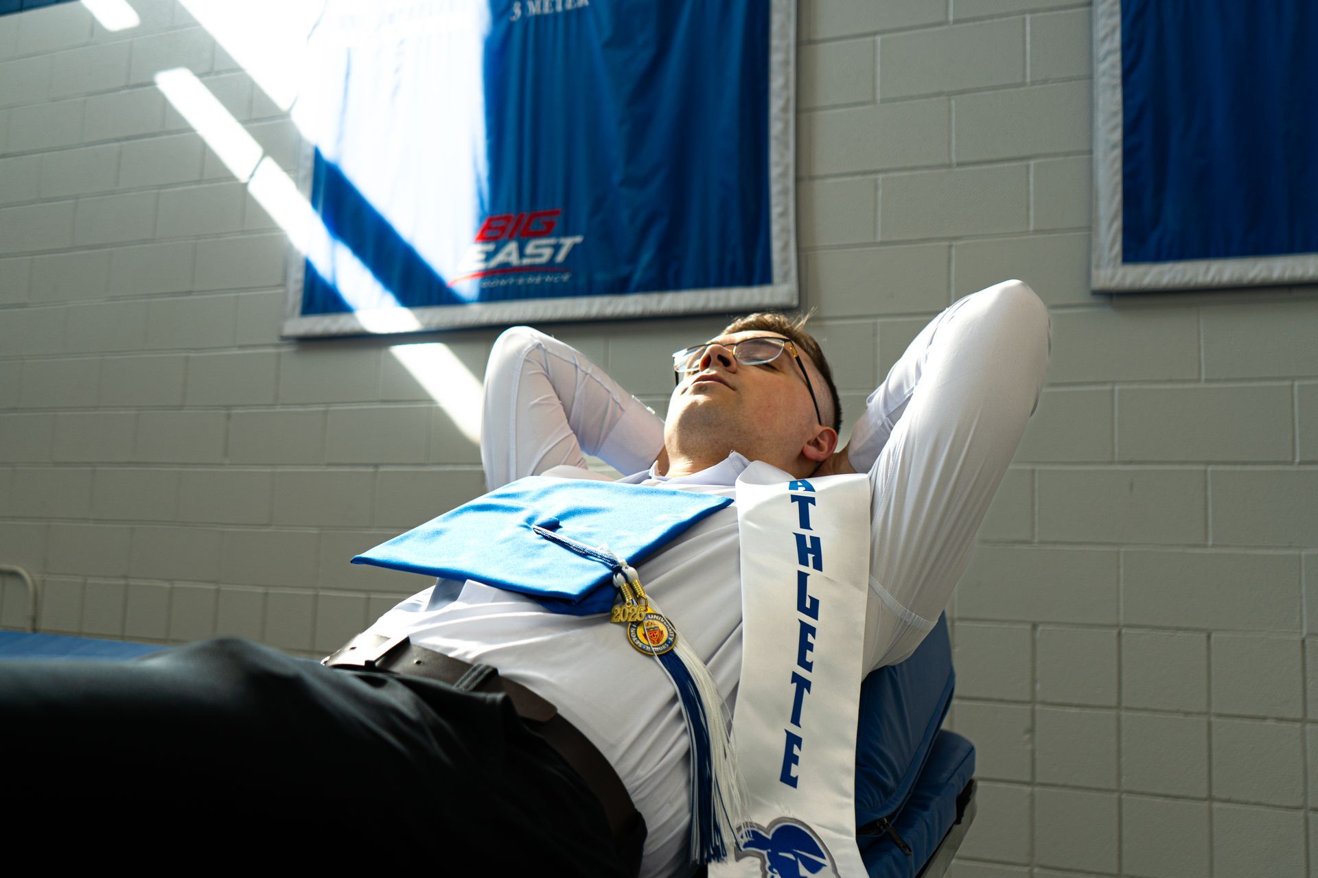 Person reclining on a chair indoors, wearing a blue vest and white sleeves, looking upward with hands behind head.