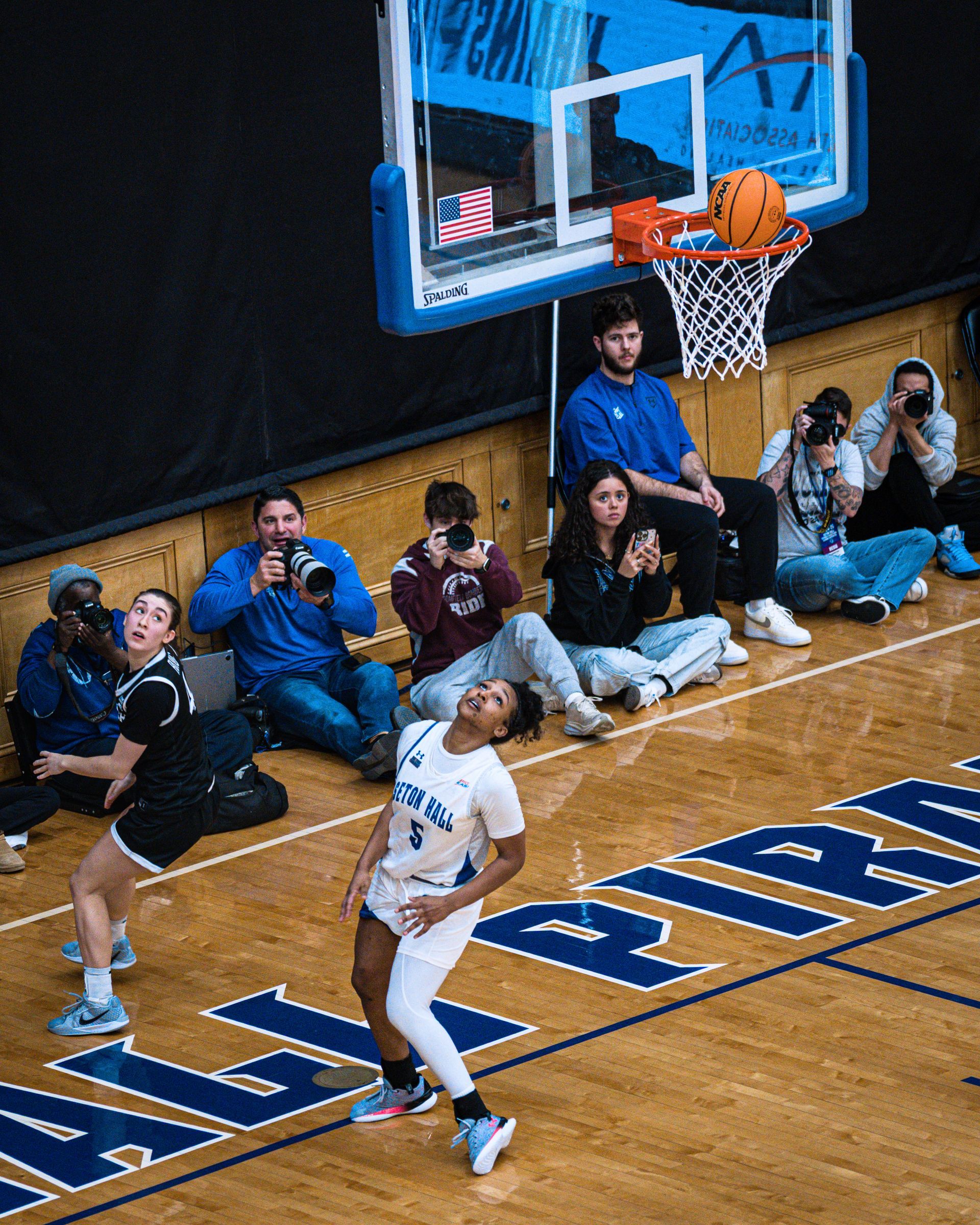 Basketball player in white jersey jumping for a layup under the hoop with photographers seated courtside.