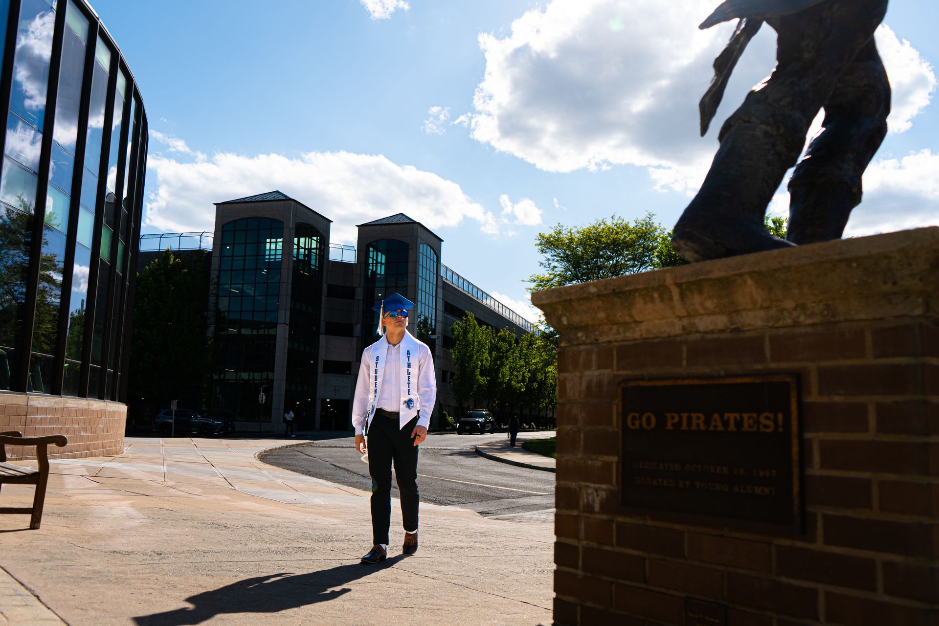 Person in a white shirt and blue cap stands on a sunny campus path near buildings and a statue.