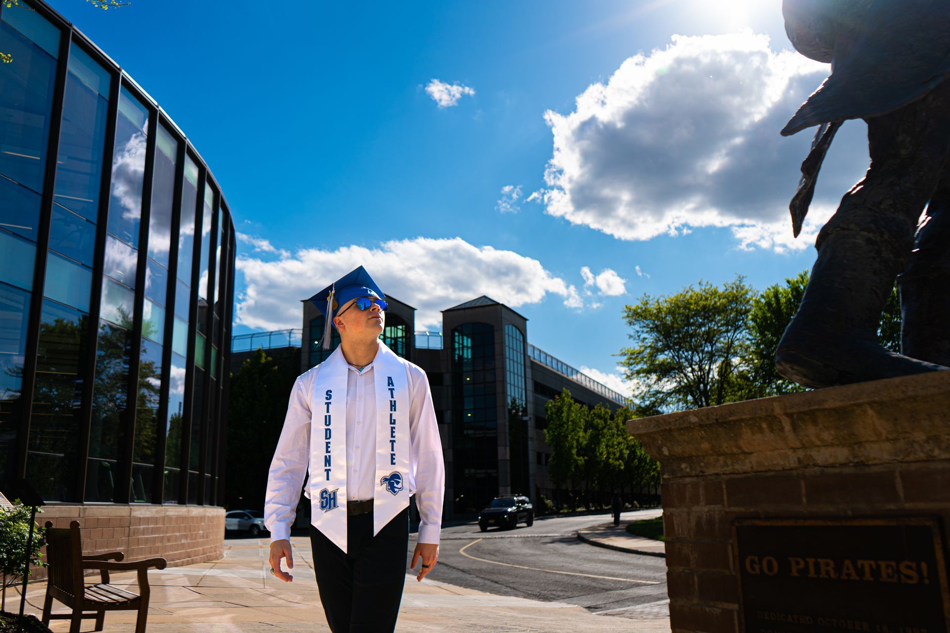Graduate in cap and gown walking on a sunny campus with modern glass buildings and blue sky