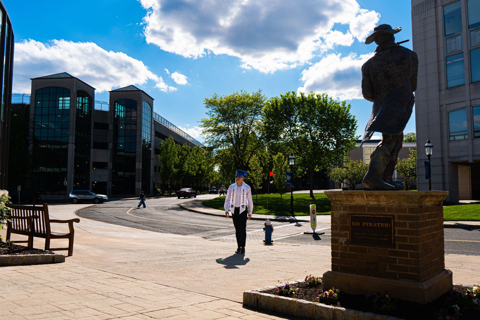Campus plaza with a statue, trees, benches, and a person walking under a bright blue sky