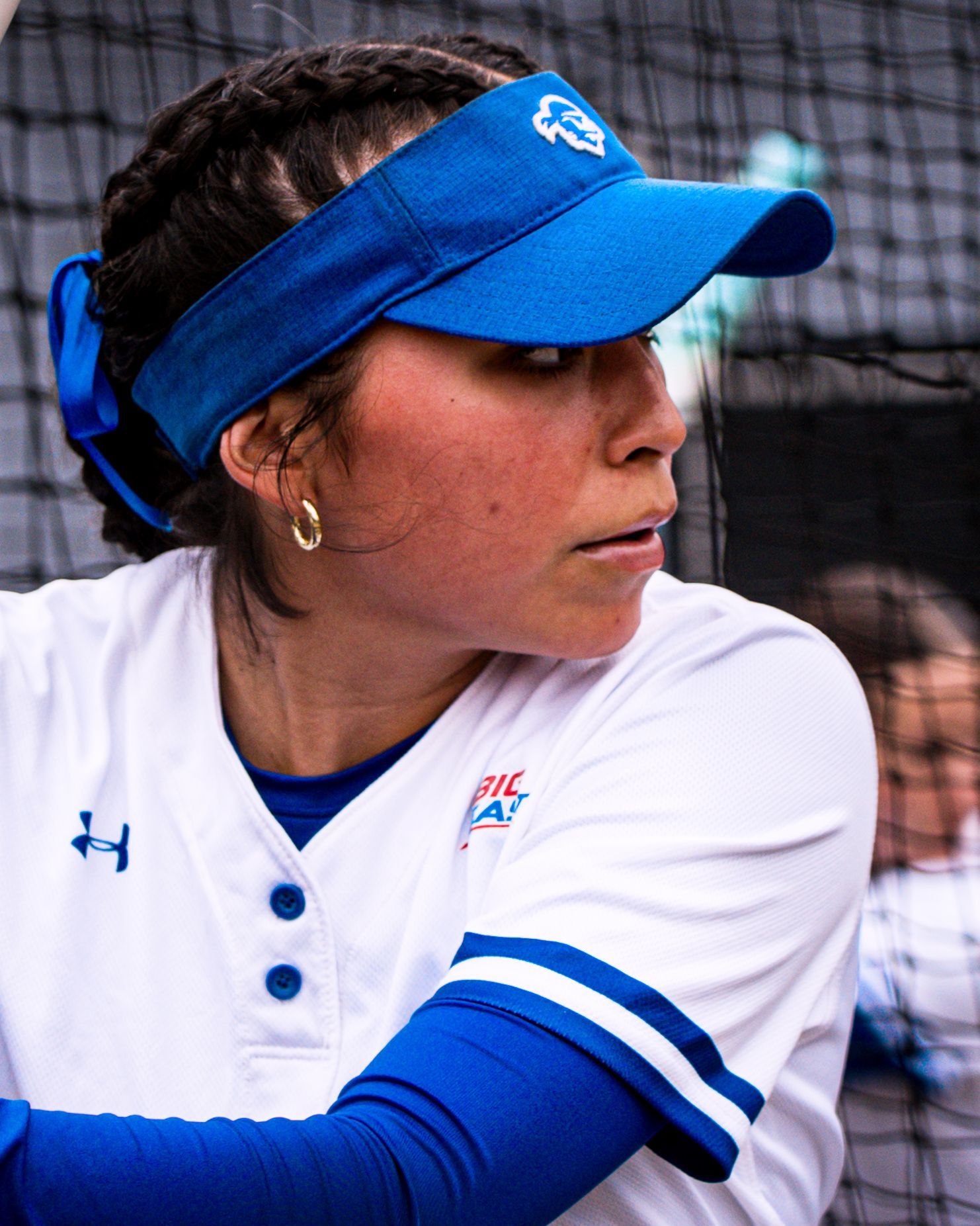 Softball player in blue visor and white jersey, holding a bat in a batting stance against a netted background