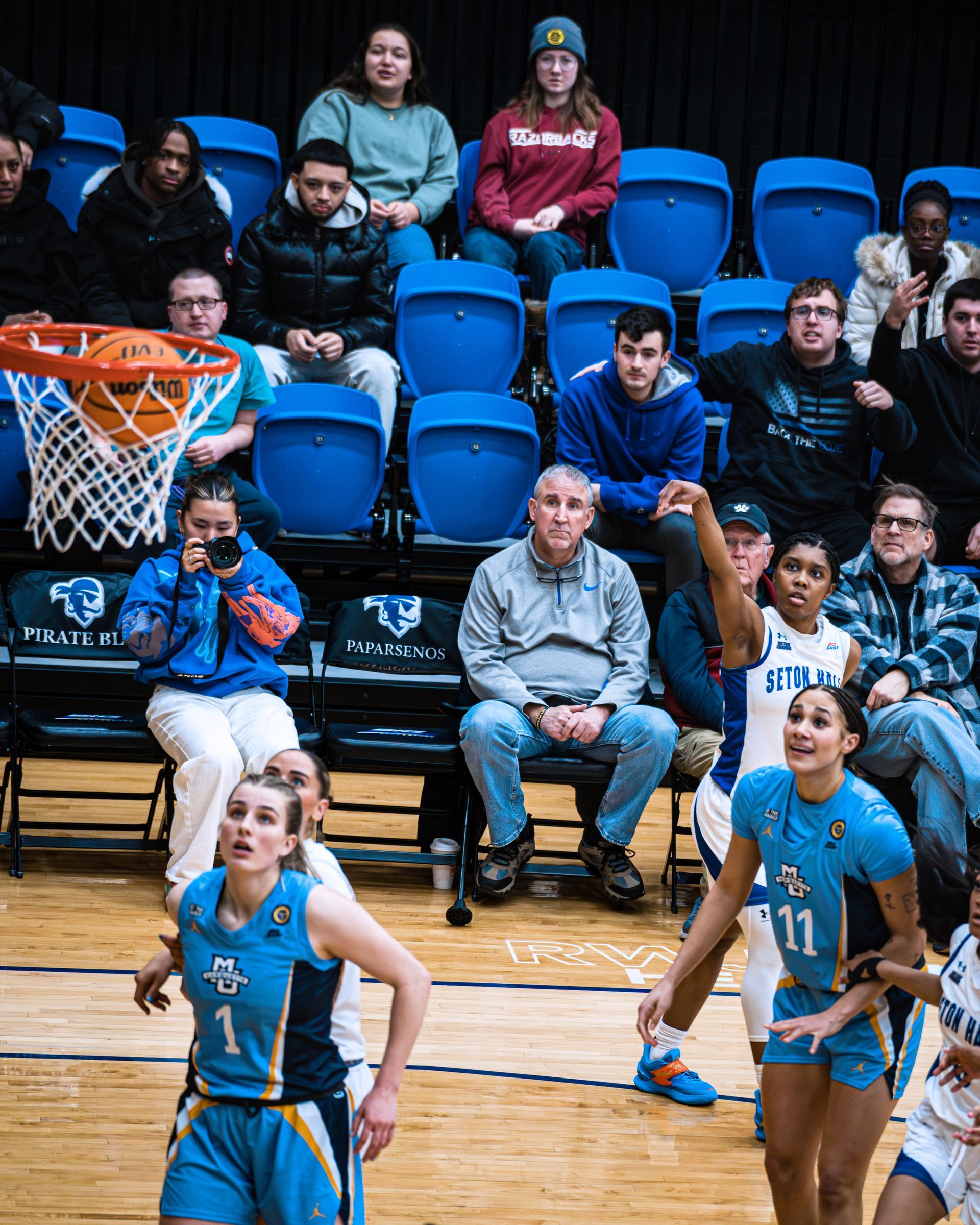 Basketball players in blue jerseys warming up on a court with spectators seated behind them.