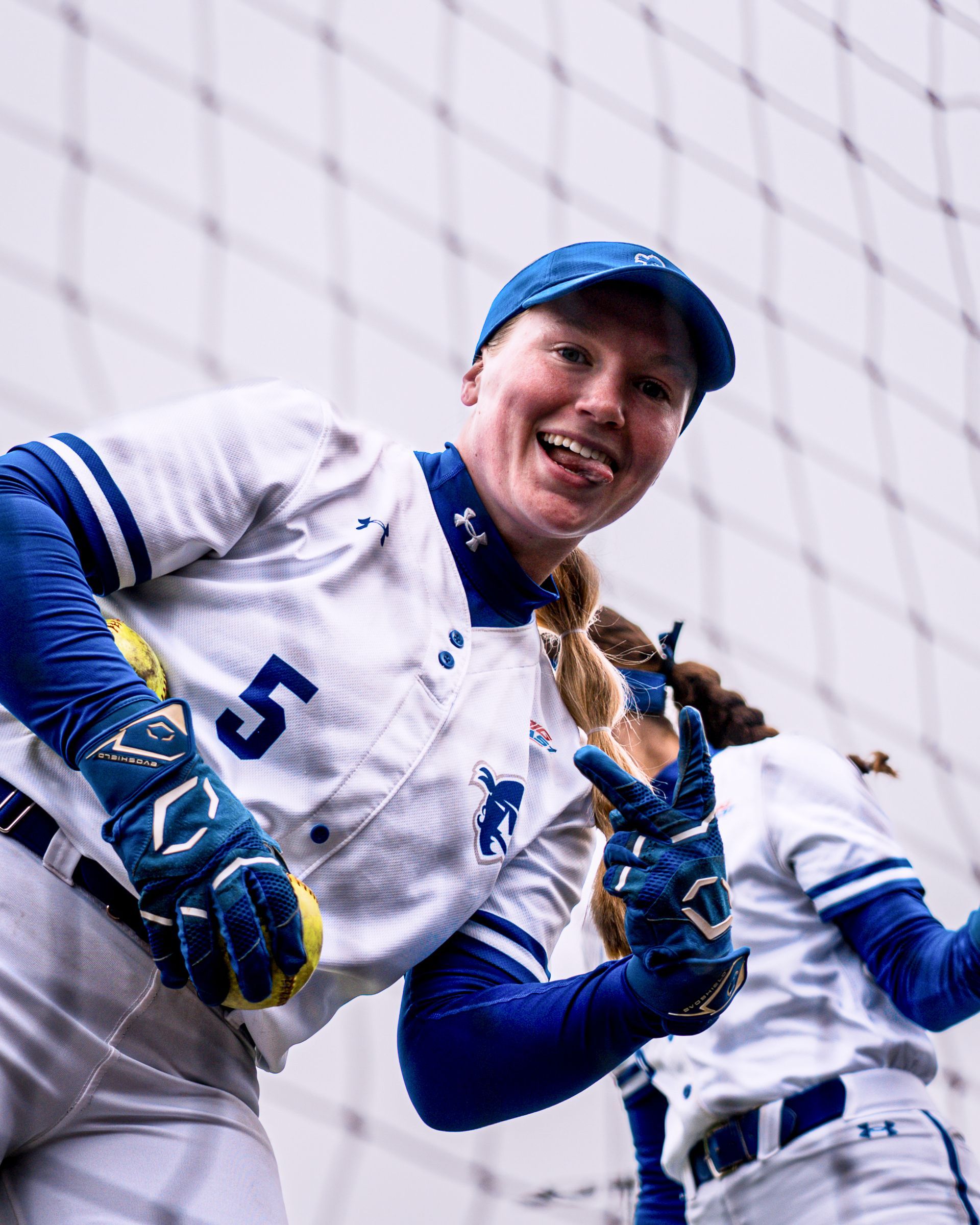 Baseball player in white and blue gear smiling, holding a glove on the field near a netting backdrop