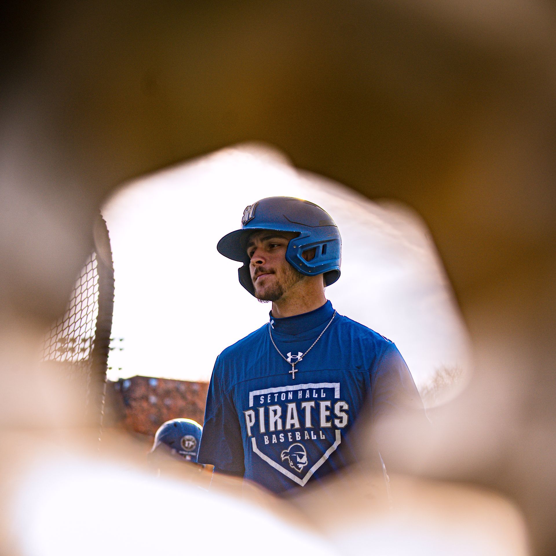 Baseball player in blue Pirates shirt and helmet standing in a dugout, seen through a blurred foreground opening