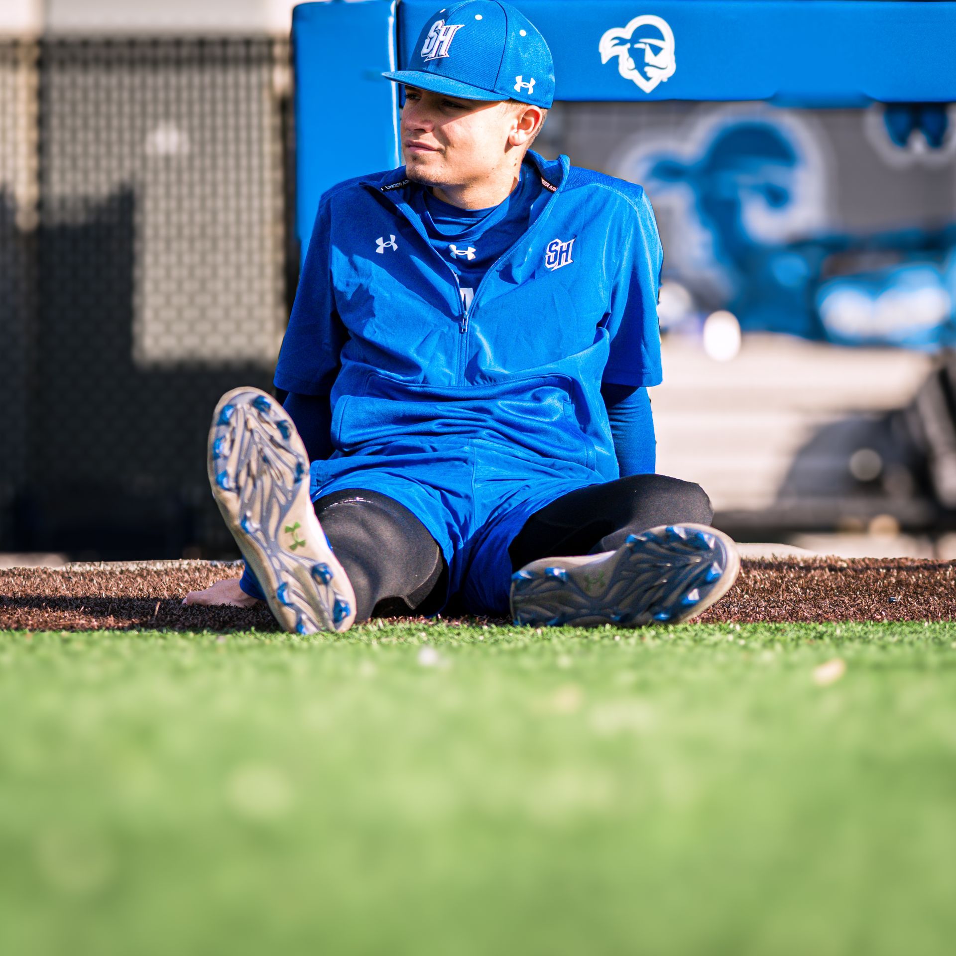 Young athlete in blue uniform and cap sits on turf near a baseball dugout, resting with knees bent.