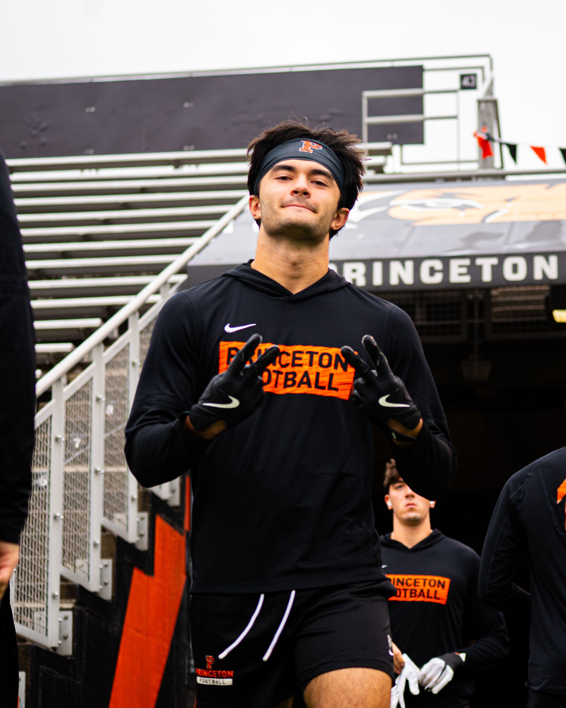 Princeton football player in black warmups runs onto the field, with stadium bleachers behind him.