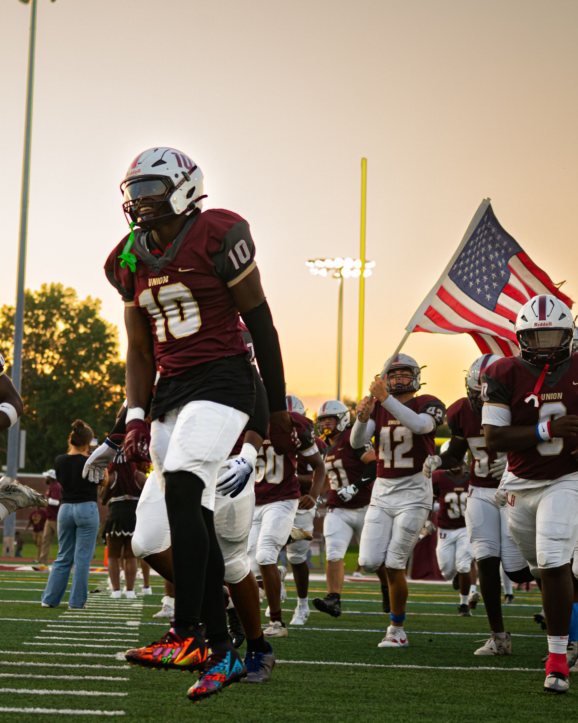 Football players in maroon uniforms run onto the field with an American flag at dusk.