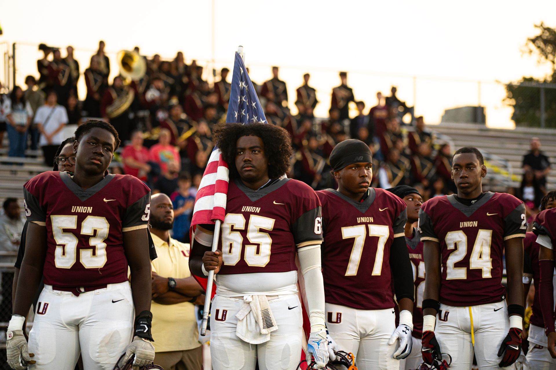 Football players in maroon jerseys stand on the field before a packed stadium crowd.