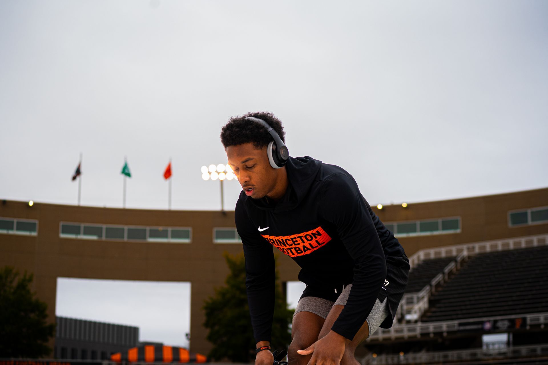 Athlete in black hoodie and headphones crouches on a track, preparing to start at a stadium