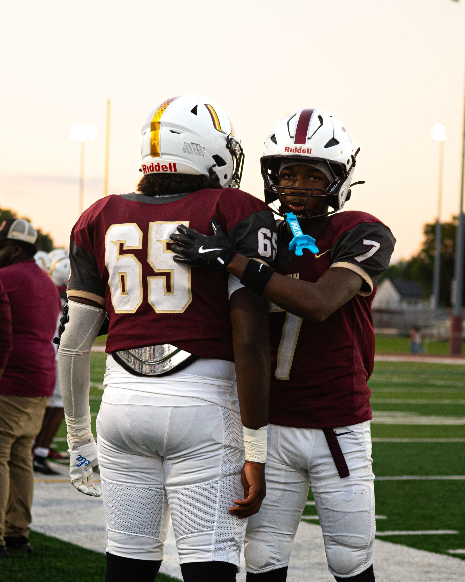 Two football players in maroon and white uniforms standing together on a field at sunset.