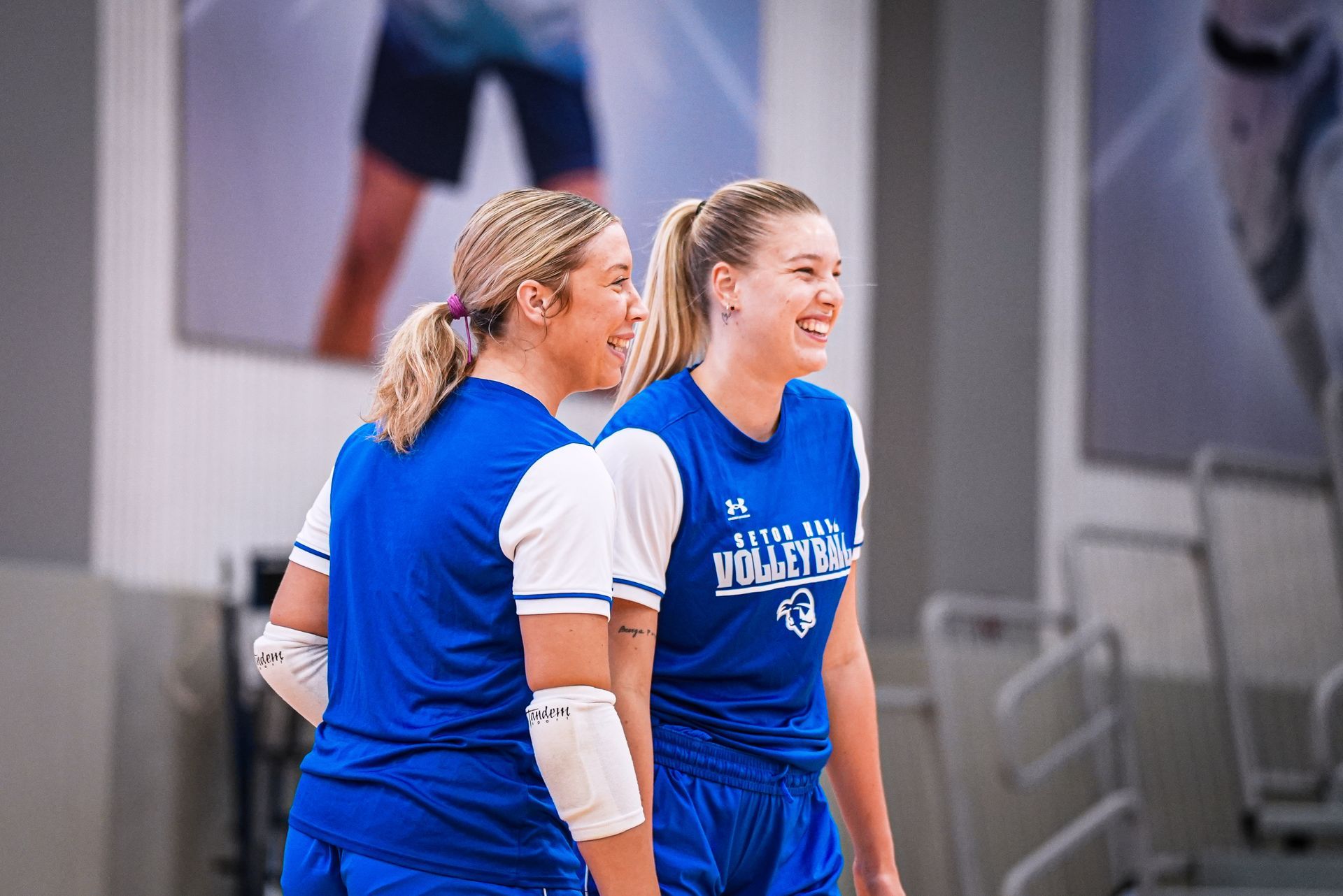 Two athletes in blue uniforms standing together indoors, one smiling and wearing a white arm sleeve