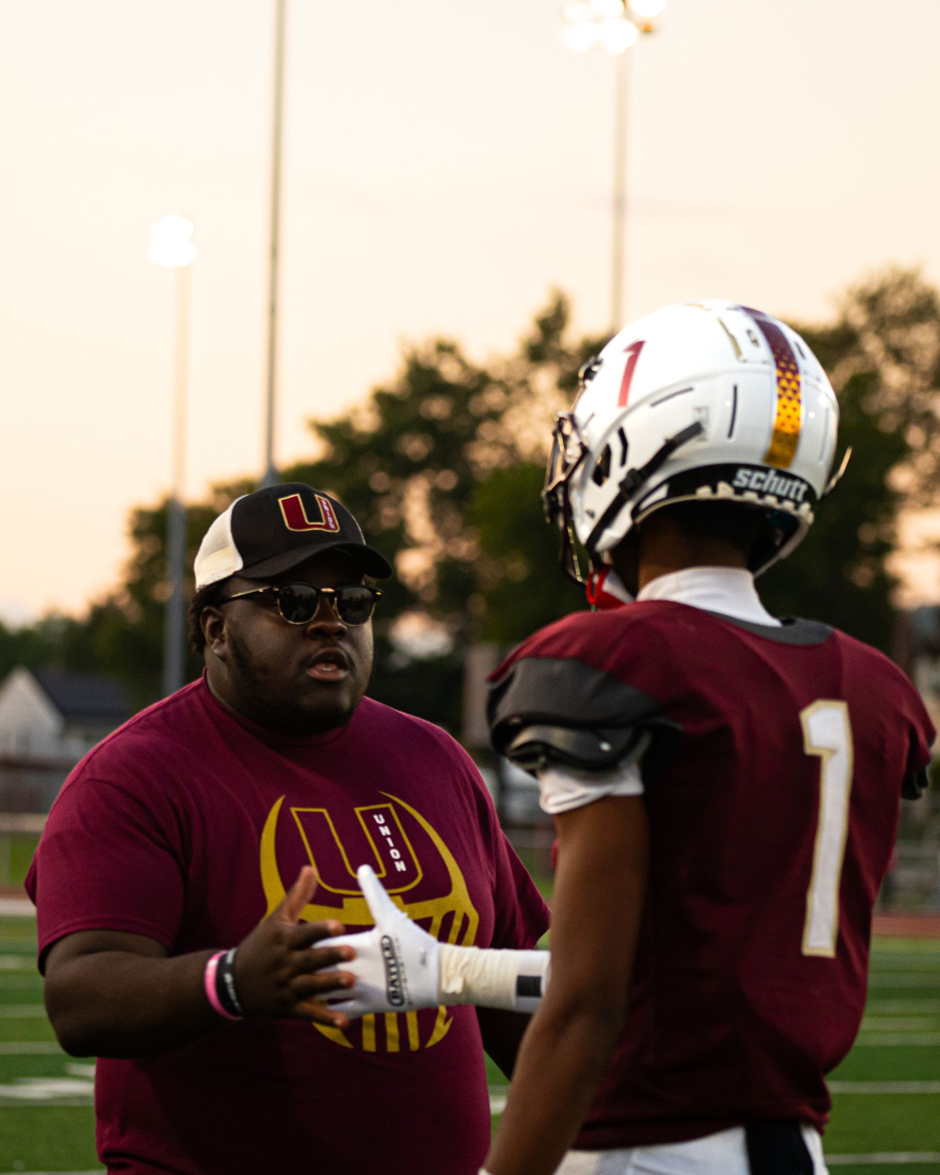 Two football players in maroon jerseys talk on a field at sunset, one holding a football.