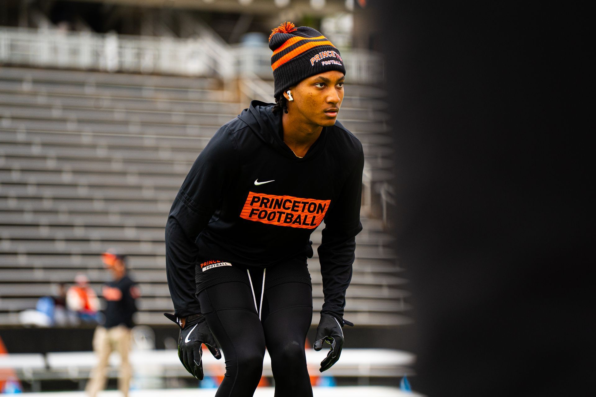Athlete in black warm-up gear and striped beanie crouches on a track stadium sideline