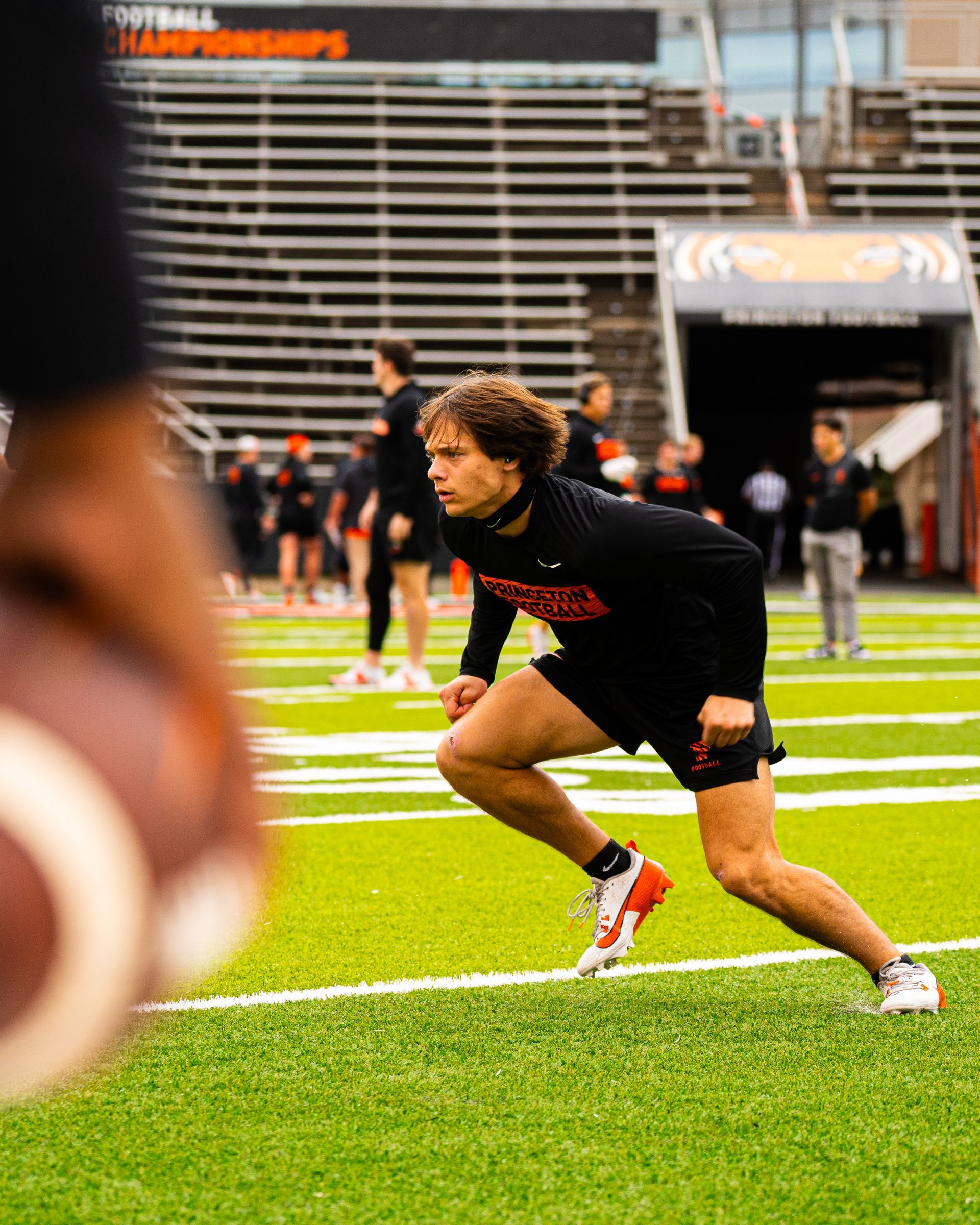 Football player in black runs low on a green field during practice, with stadium bleachers in the background