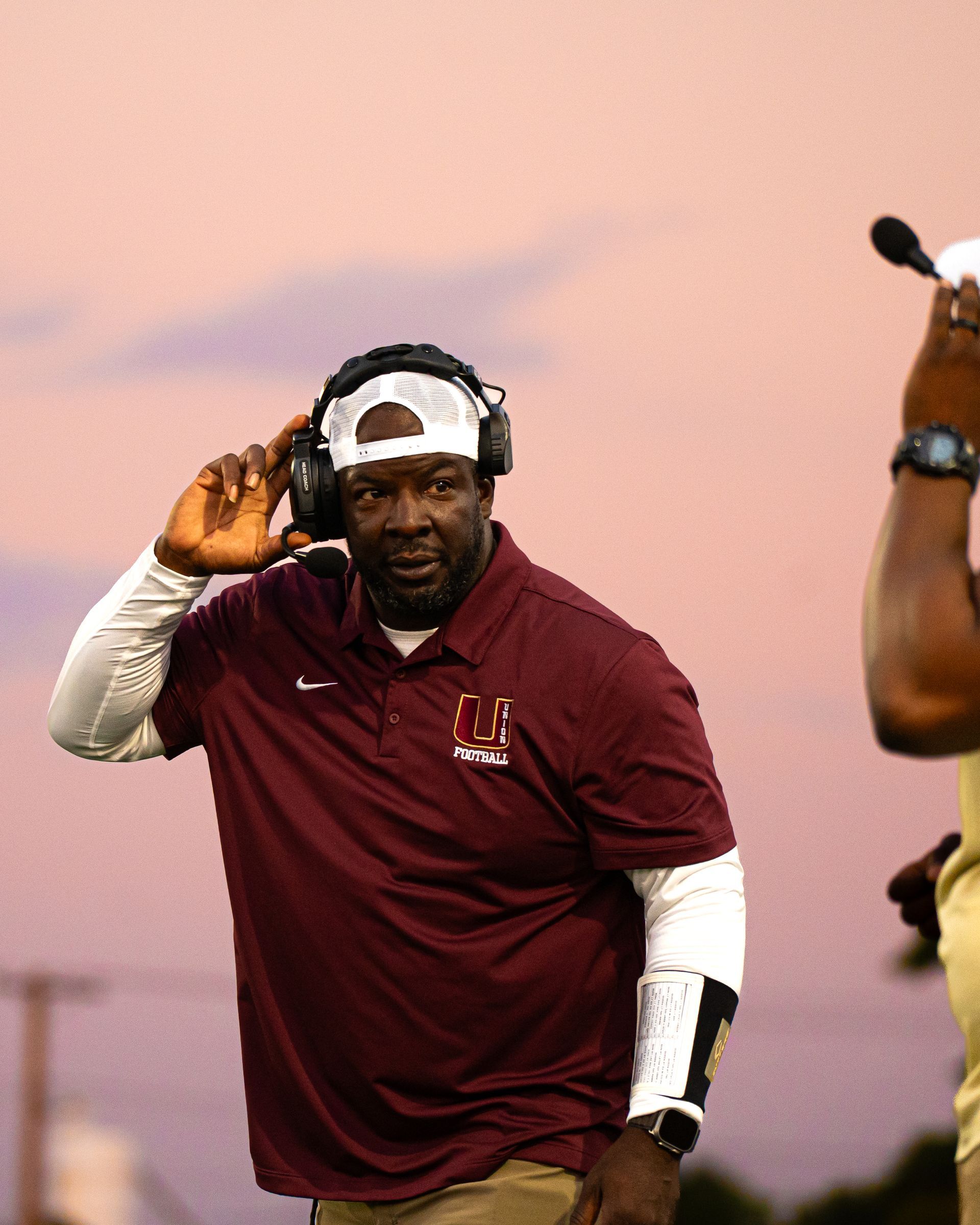 Man in maroon shirt and white cap listening on a headset against a sunset sky.